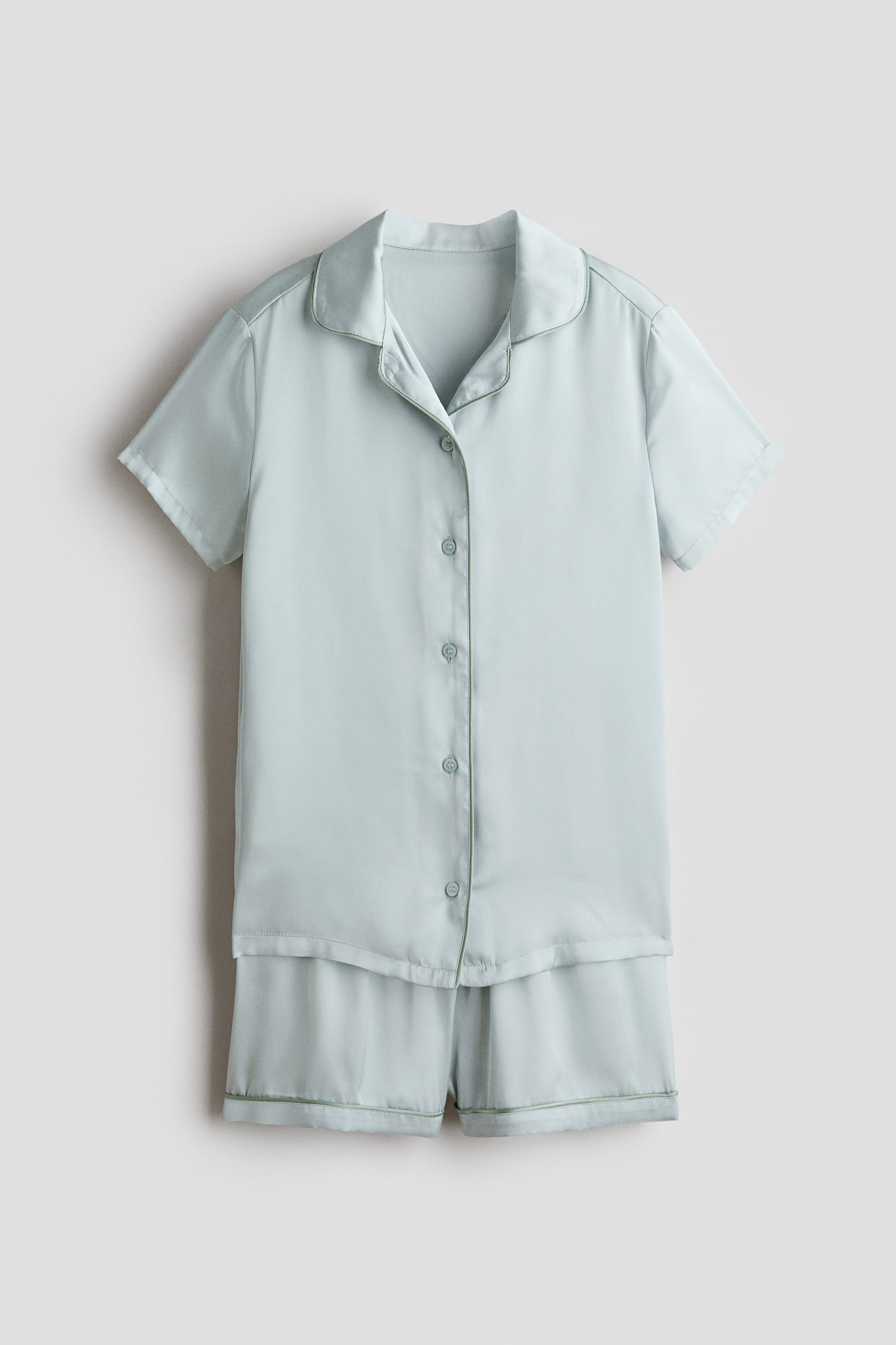 View larger image: A light blue-green pyjama set is presenting its front view, featuring a short-sleeved shirt and coordinating shorts made from a smooth, slightly lustrous fabric. The shirt is showcasing a collar with notch lapels, a button-down front with six matching buttons, and contrasting green piping outlining the collar, button placket, and sleeve hems, while the matching shorts are also displaying green piping along their hems.