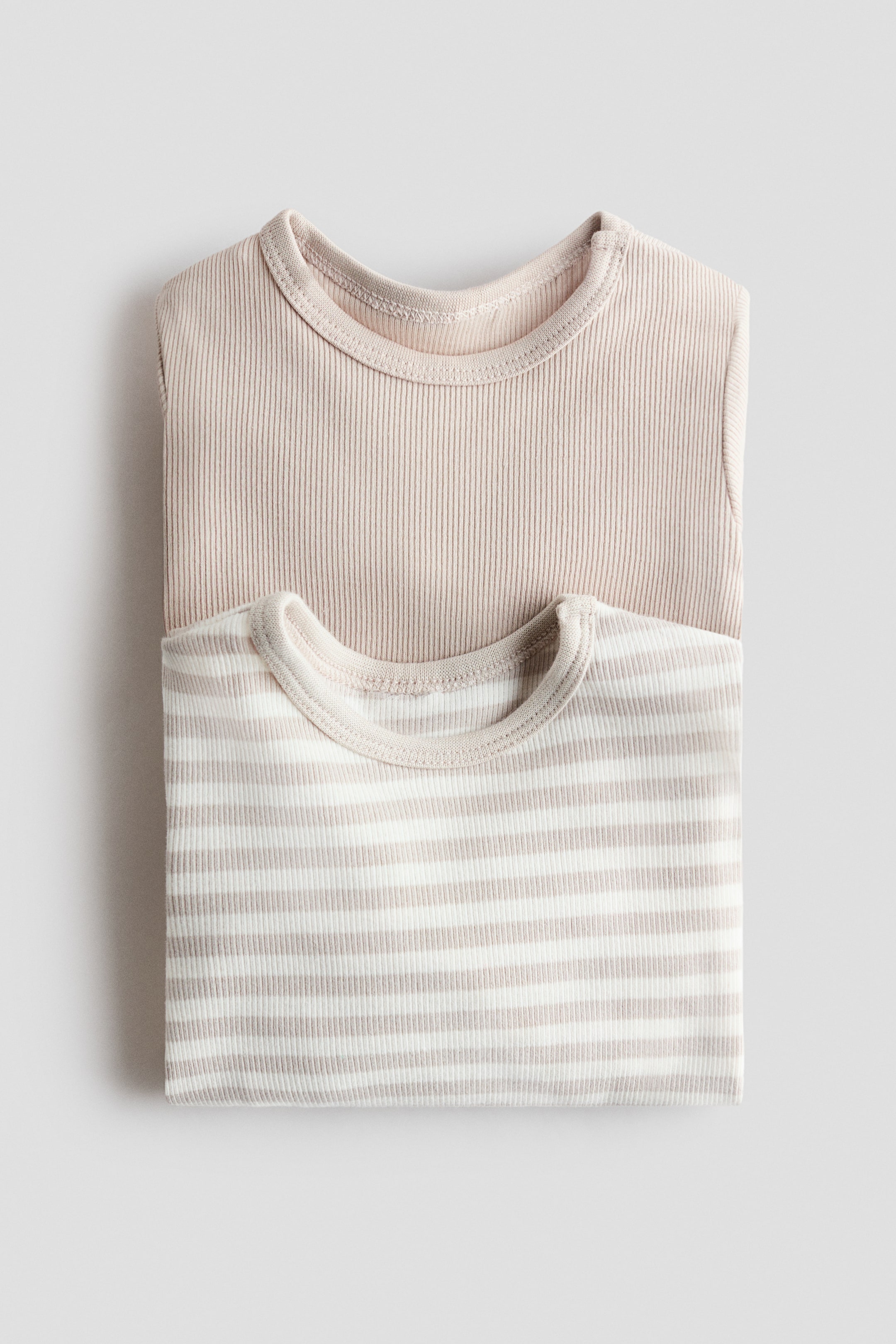 Ver imagem maior: Dois bodysuits dobrados de bebé estão deitados planos, apresentados de uma vista de cima para baixo. O bodysuit de cima exibe uma textura canelada bege claro sólida e uma gola redonda, enquanto o bodysuit inferior parcialmente visível apresenta um padrão canelado de riscas bege claro e branco e uma gola redonda semelhante.
