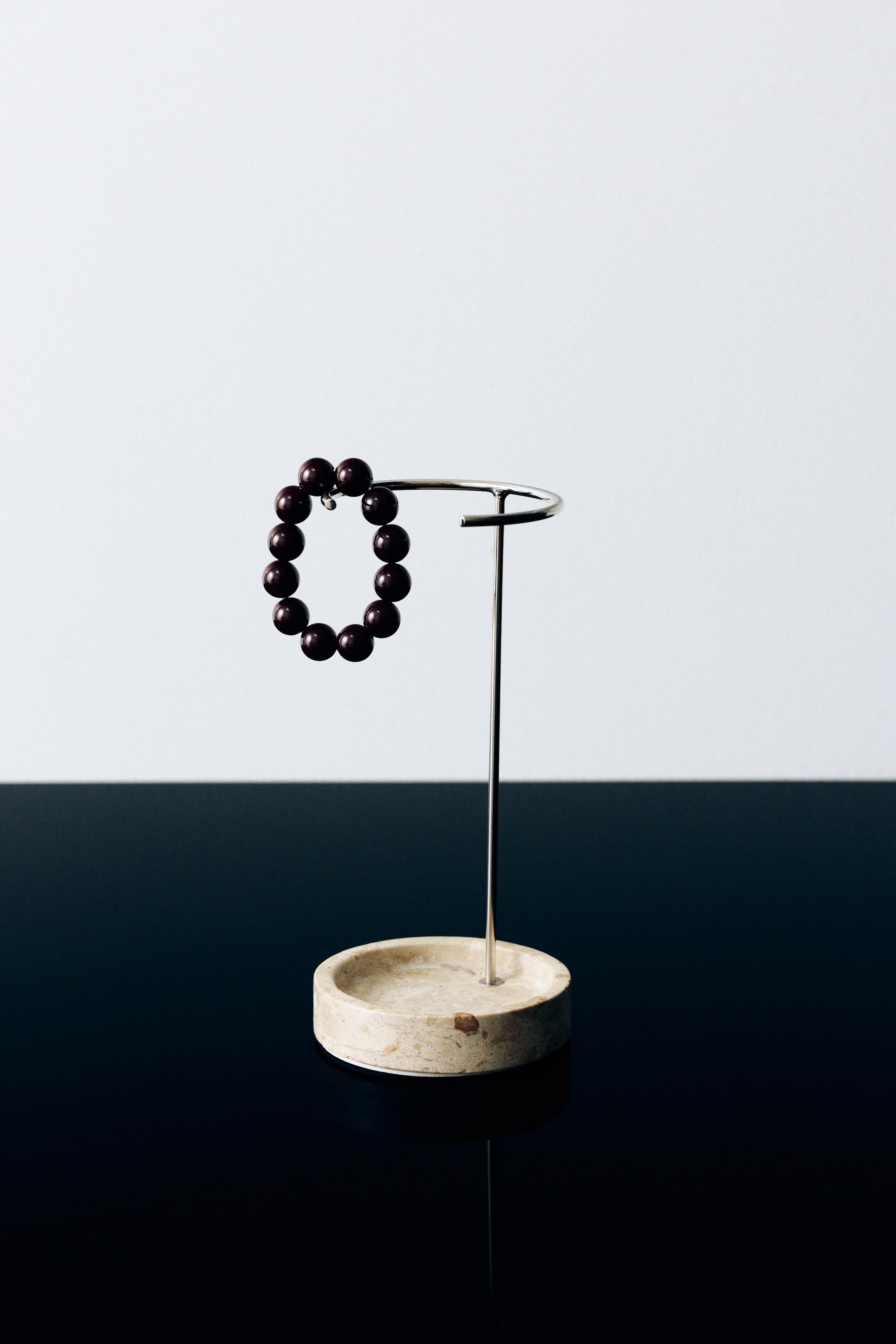 Visualizza l'immagine ingrandita: Un espositore per gioielli è in posizione verticale, con una base a vassoio rotonda in marmo beige e una sottile struttura in metallo color argento con la parte superiore curva. Un bracciale di perline marrone scuro è appeso a un'estremità della barra metallica curva.