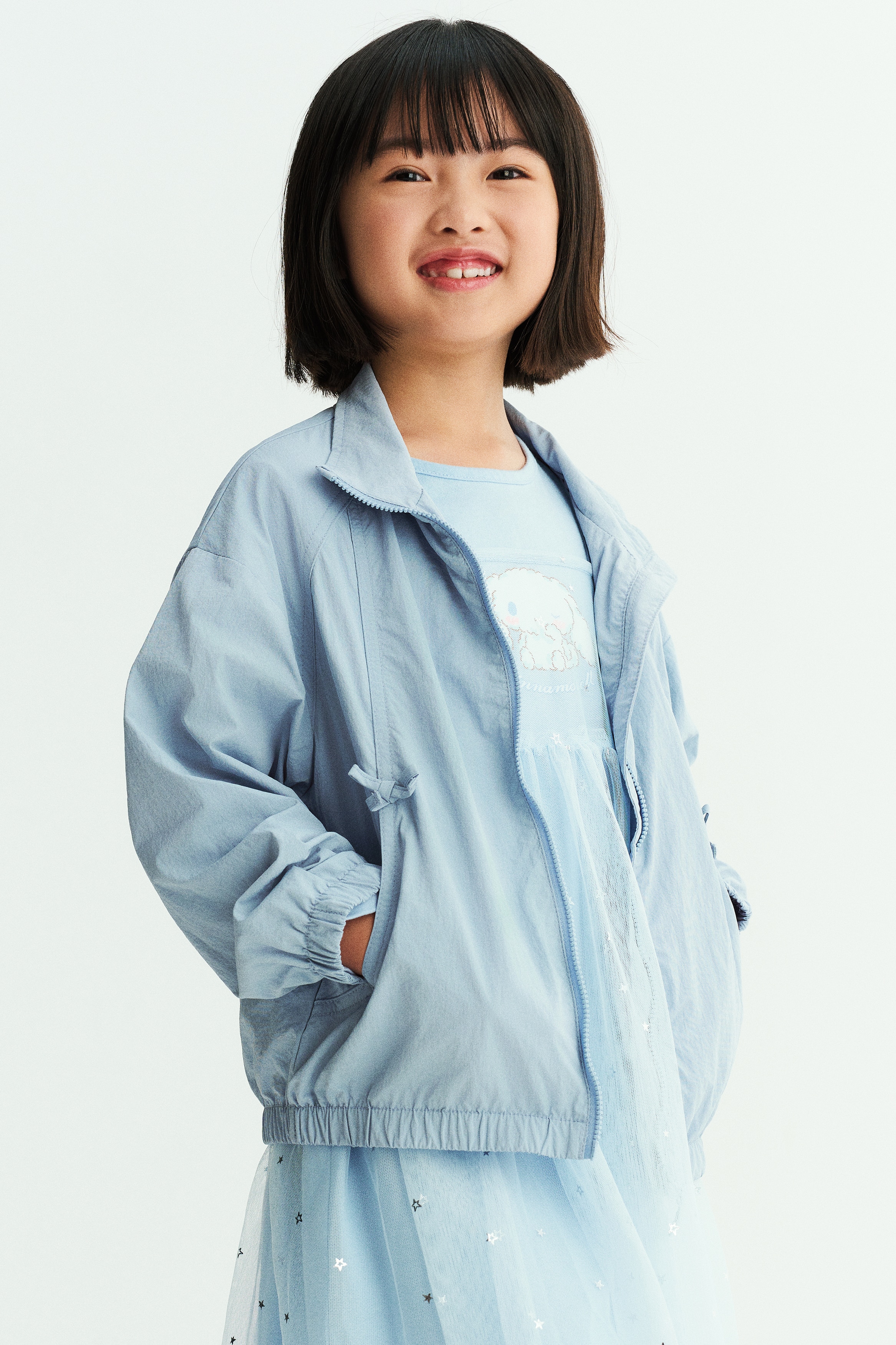 Má»t chiáº¿c Ã¡o khoÃ¡c giÃ³ báº±ng nylon mÃ u xanh dÆ°Æ¡ng nháº¡t ÄÆ°á»£c chá»¥p tá»« phÃ­a trÆ°á»c, ná»i báº­t vá»i cá» Äá»©ng cao, khÃ³a kÃ©o phÃ­a trÆ°á»c á» giá»¯a vÃ  vai rÅ© vá»i cÃ¡c ÄÆ°á»ng may cong. Ão khoÃ¡c cÃ³ cÃ¡c nÆ¡ trang trÃ­ nhá» phÃ­a trÃªn tÃºi bÃªn chÃ©o, cÃ¹ng bo chun á» cá» tay vÃ  gáº¥u Ã¡o.