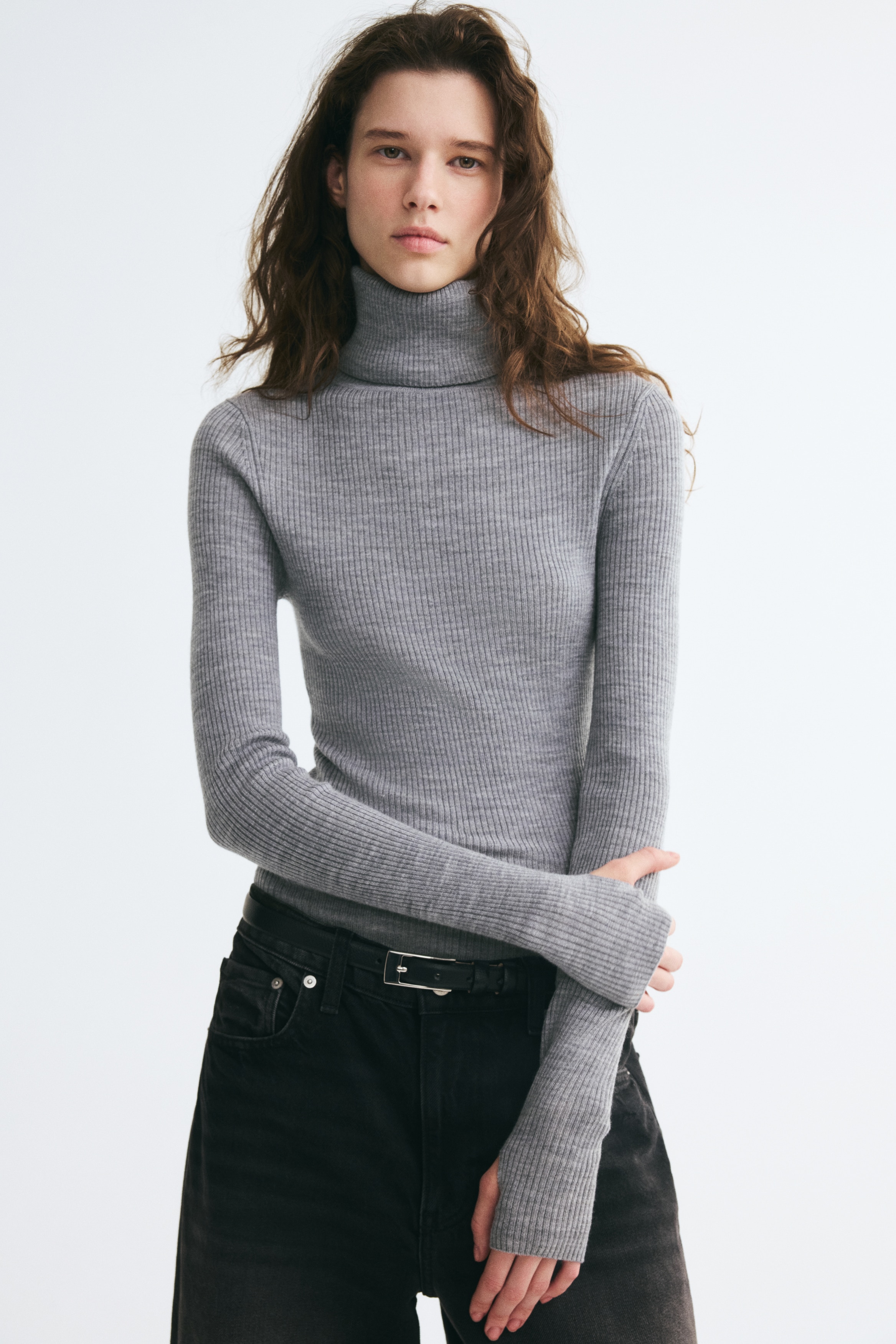 A grey, rib-knit roll-neck top is shown from the front, highlighting its fitted silhouette and vertical ribbed texture. The long sleeves extend downwards, featuring visible integrated thumbholes near the cuffs.