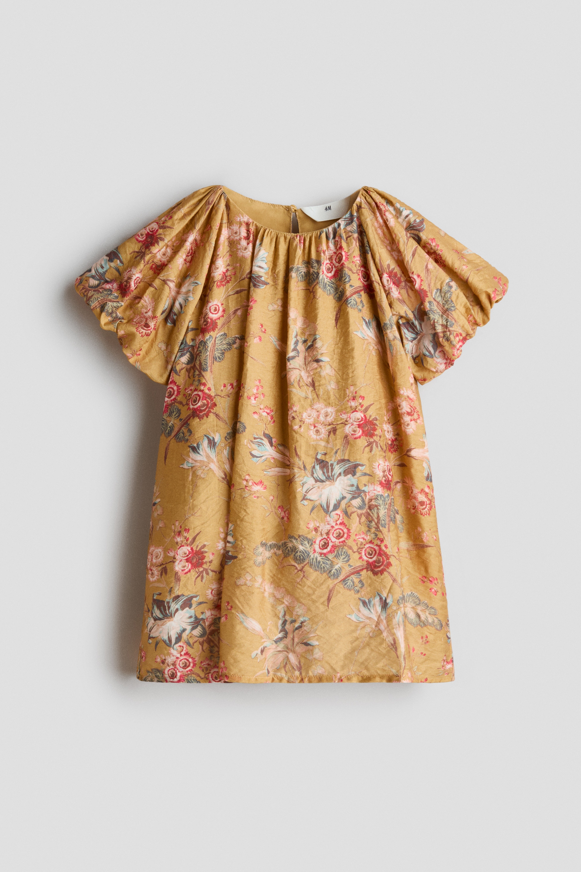 A child is wearing a mustard-yellow, floral-patterned A-line dress and is facing forwards, showcasing its gathered round neckline and short, voluminous raglan sleeves with elasticated cuffs.