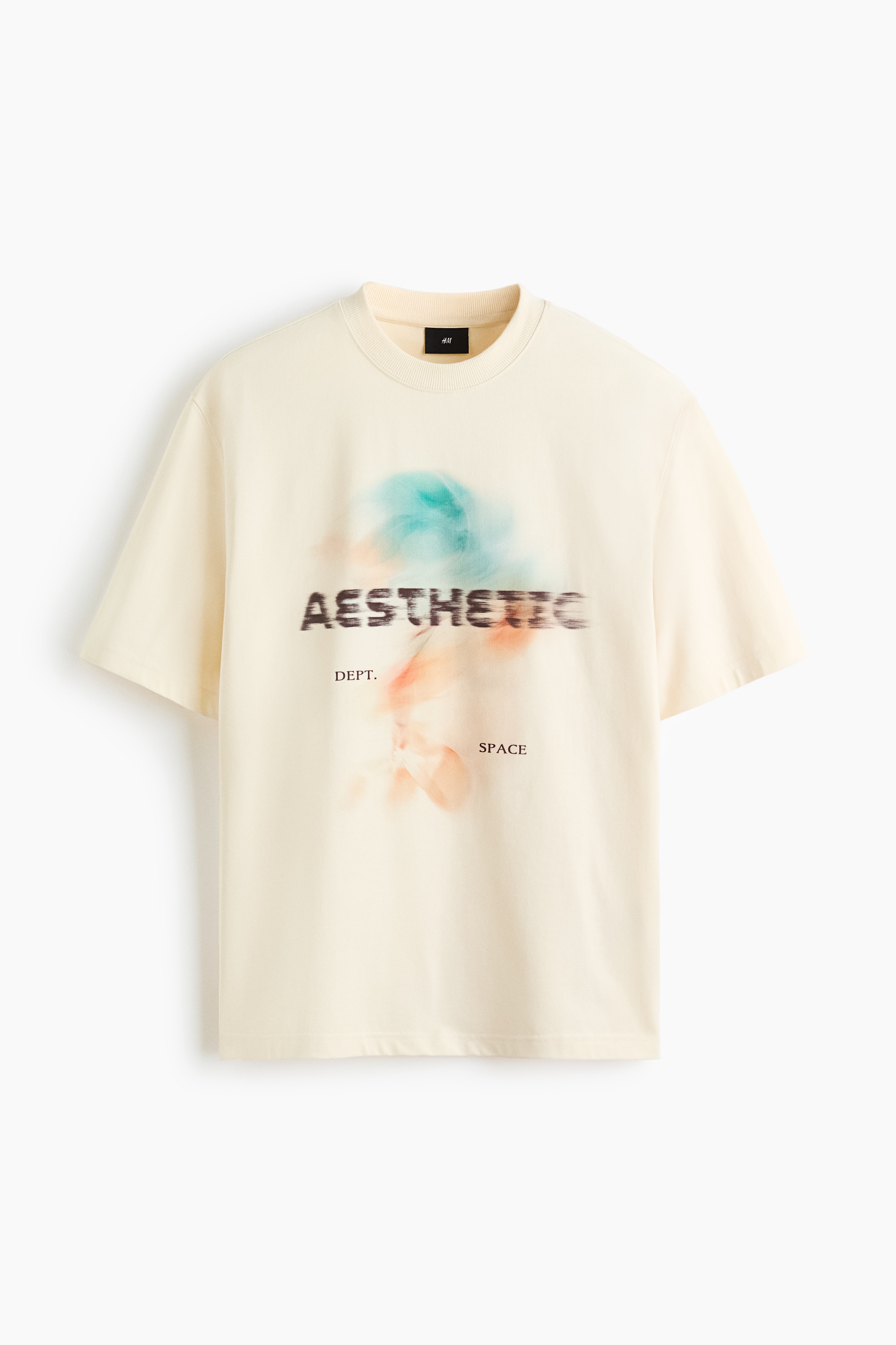 A boxy-style cream T-shirt is worn by the model, featuring a round rib-trimmed neckline, dropped shoulders, and a blurred abstract graphic print with text on the front. The loose-fit T-shirt is paired with dark green trousers, with the model’s left hand resting in a pocket.