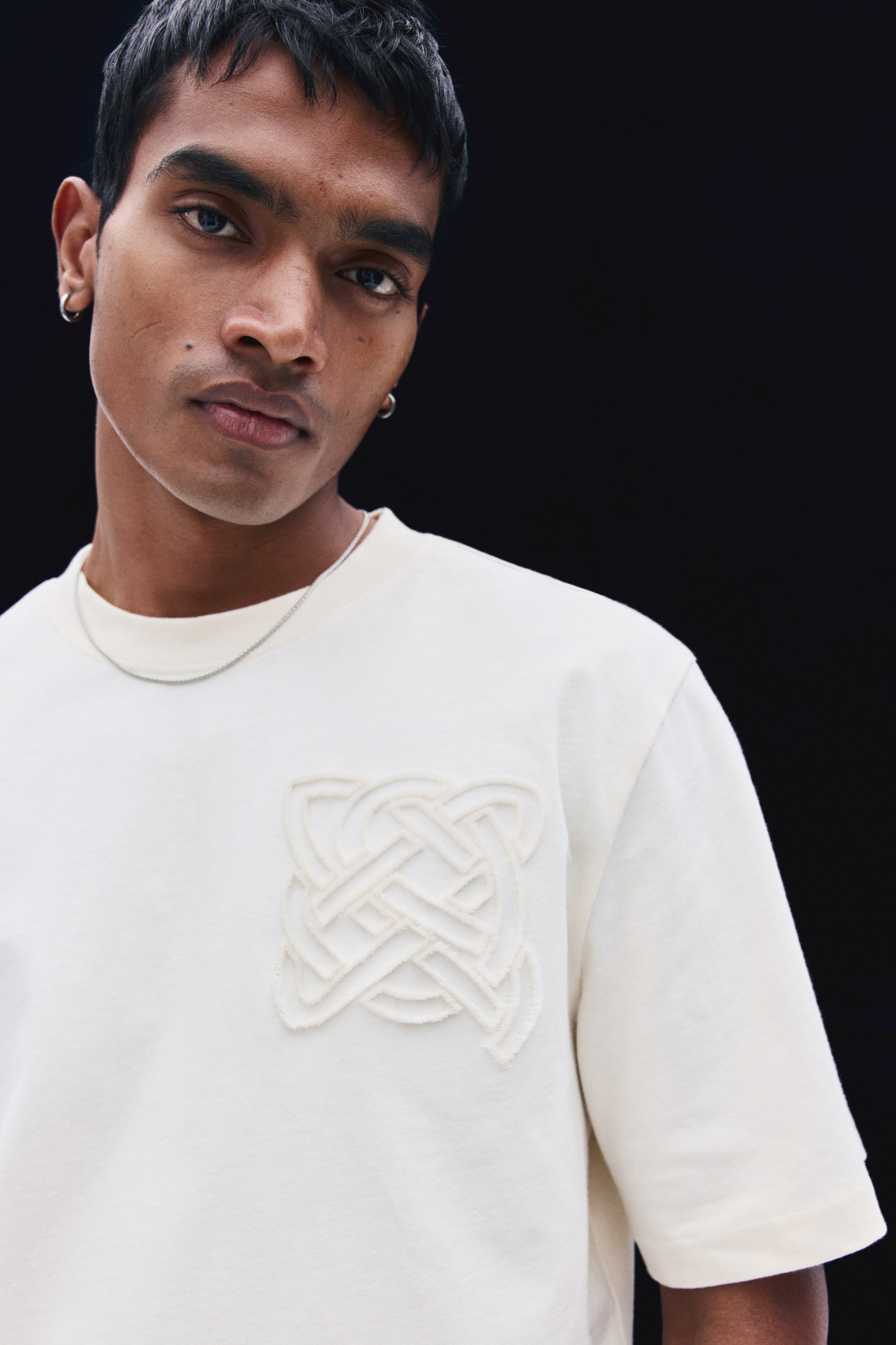 View larger image: An off-white, midweight cotton T-shirt featuring a round neckline and a raised, textured geometric knot design on the chest is being worn by the model. A thin silver chain necklace is layered over the ribbed collar of the tee.