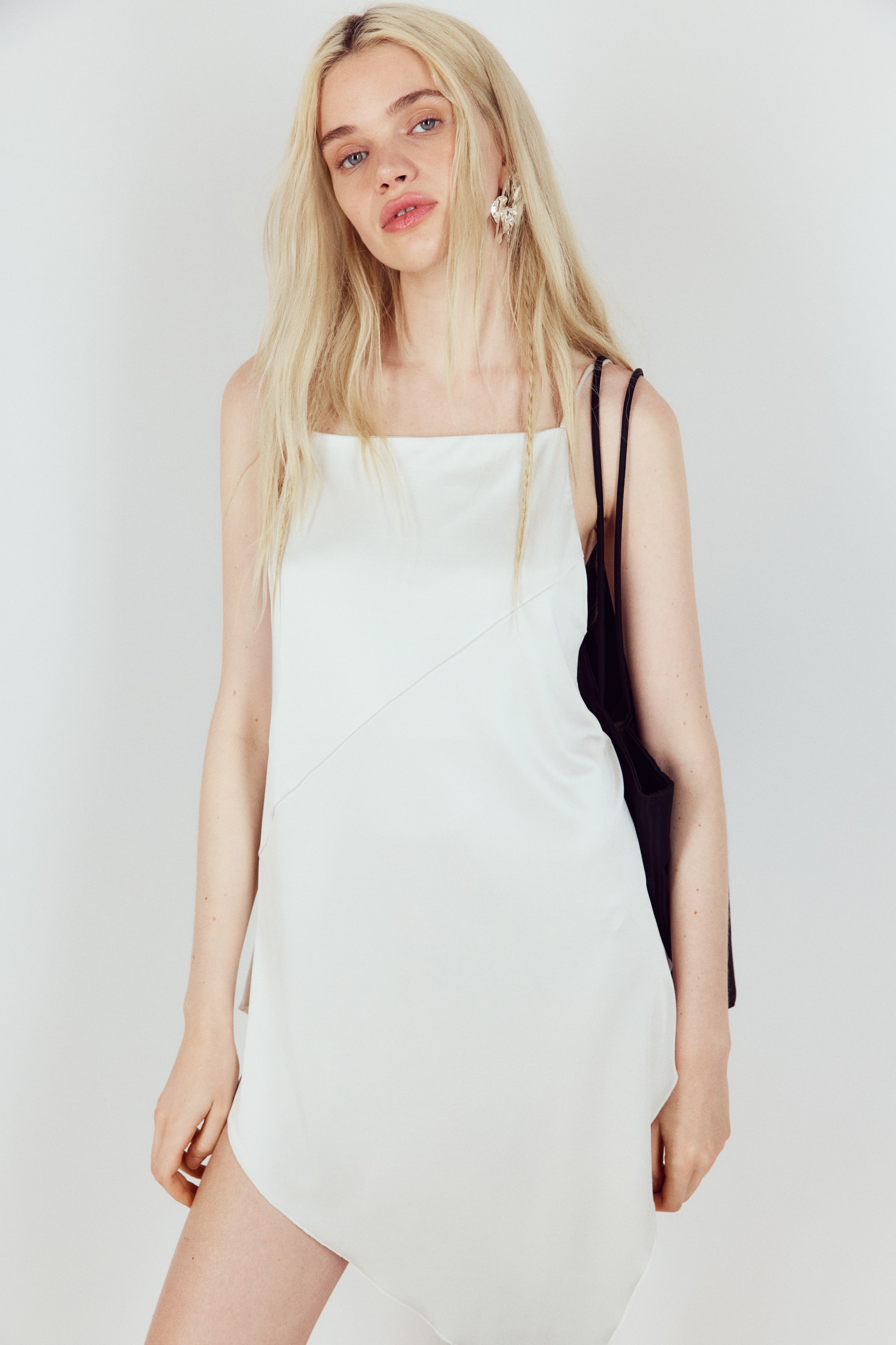 Agrandir l'image: Un haut blanc long et ample avec une finition lisse, doté d'un décolleté droit, de fines bretelles et d'une couture diagonale à l'avant, est porté par le mannequin. Il présente un ourlet asymétrique en forme de V, et un sac noir est porté sur son épaule droite.