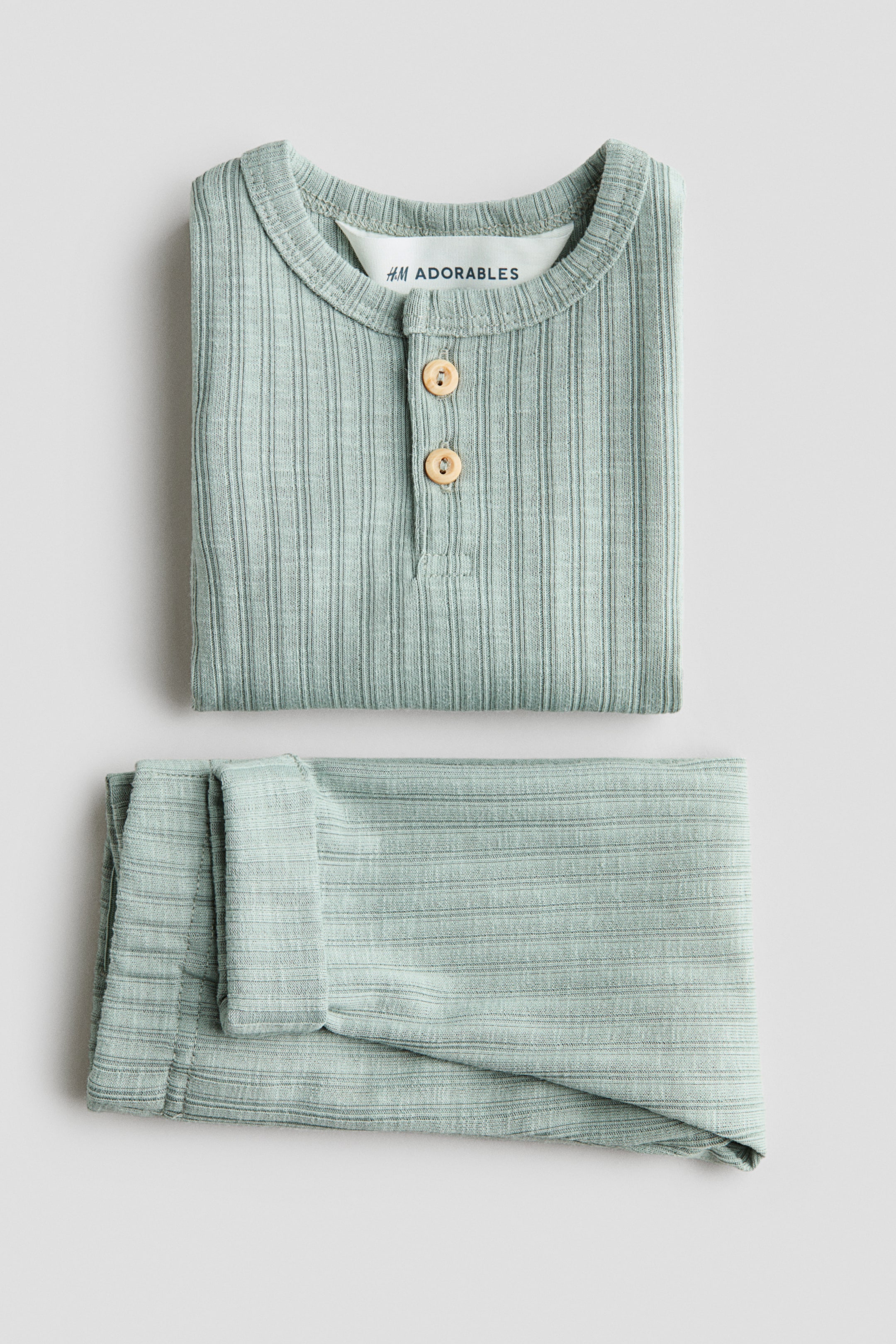 Visualizza immagine più grande: Un completo a due pezzi in maglia a coste verde chiaro è piegato ordinatamente, con la parte anteriore del top visibile. Il top presenta uno scollo rotondo con allacciatura Henley a due bottoni e un'etichetta 'H&M ADORABLES', e i pantaloni abbinati sono piegati sotto.