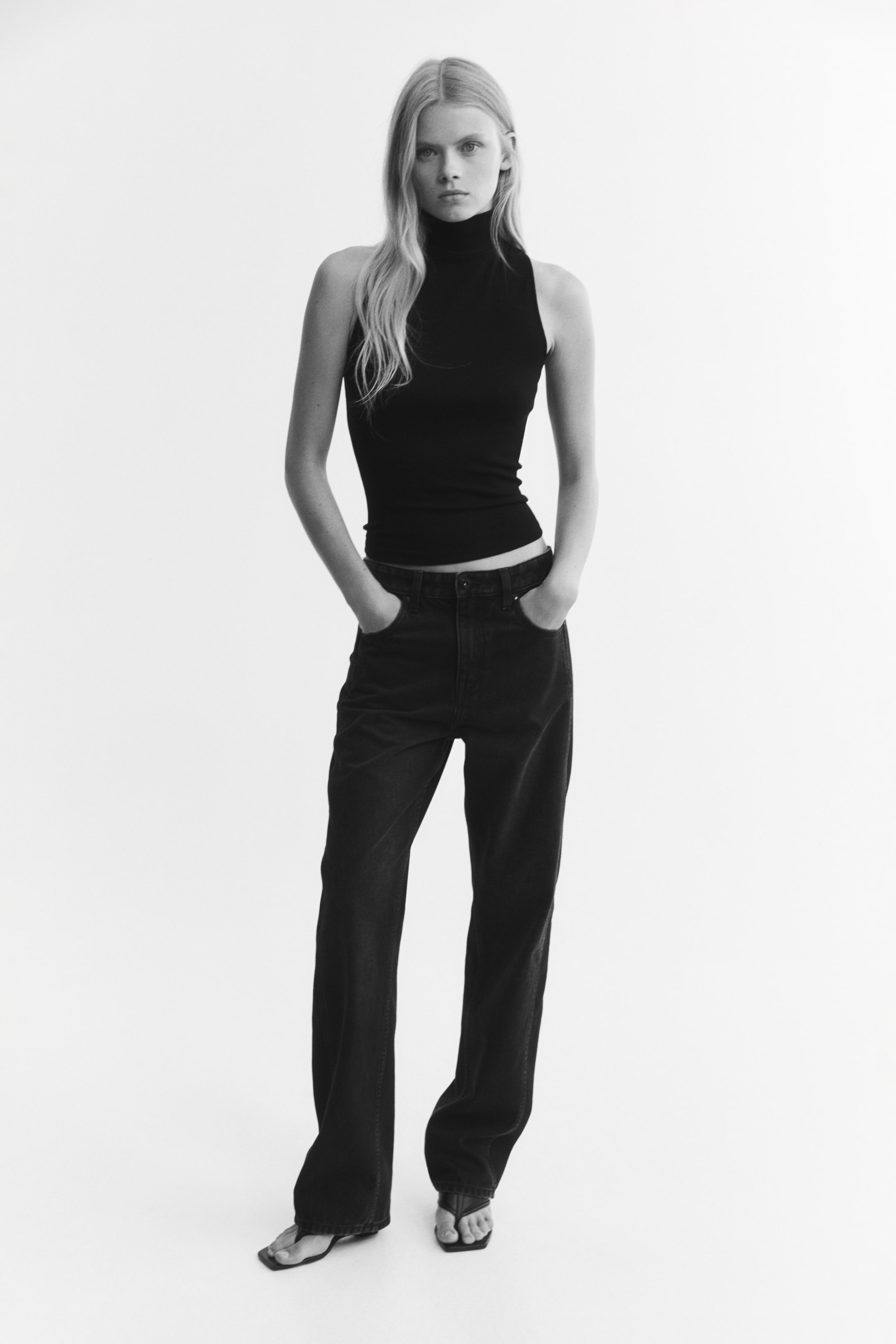 View larger image: A fitted, sleeveless black top featuring a high neck and deep armholes is worn by the model. The top is paired with dark, straight-leg jeans.