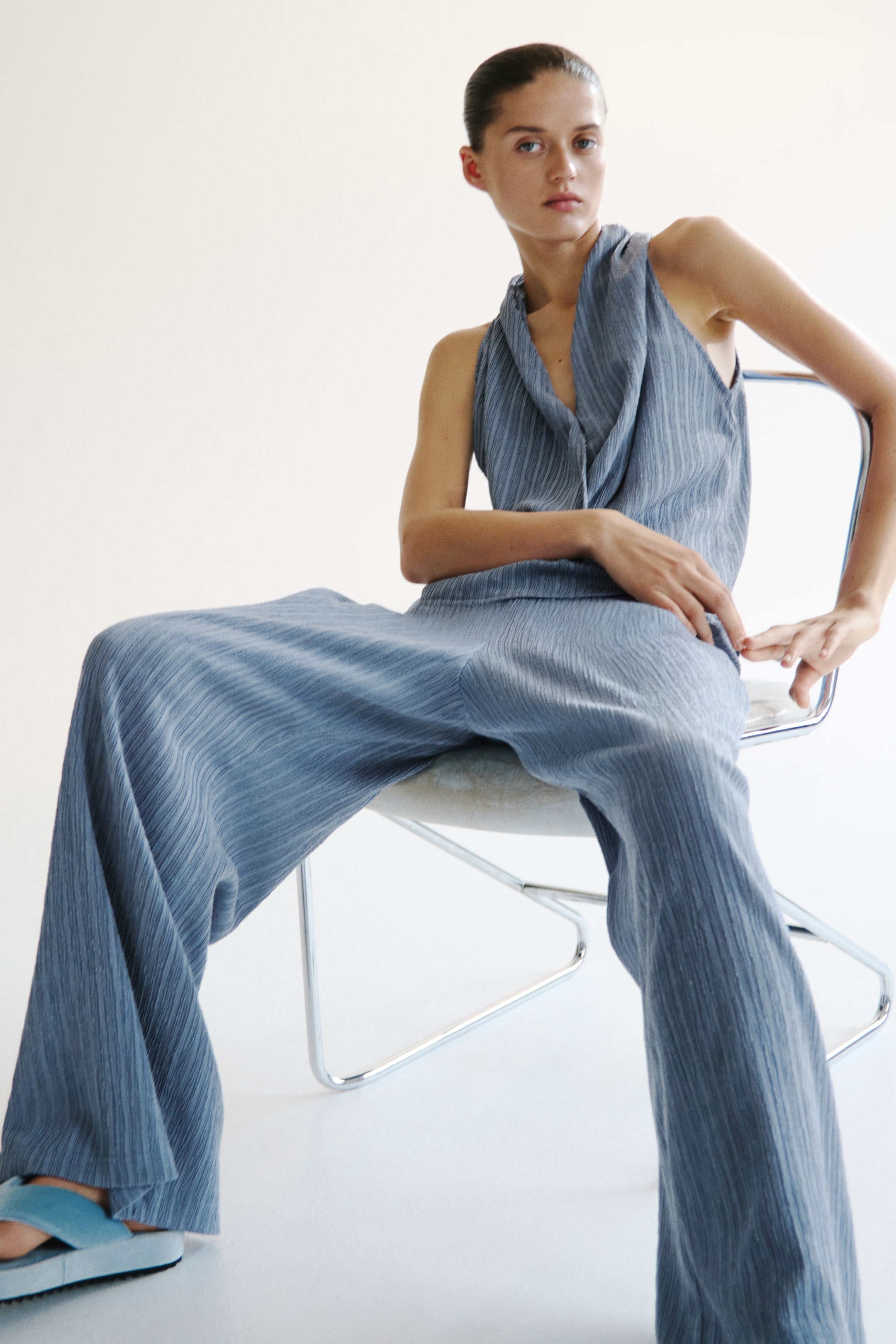 View larger image: The model is wearing a sleeveless top in blue-gray crinkled jersey, featuring a shawl collar, a narrow cut at the top, and a sewn-in wrapover at the front. She is seated, also sporting matching wide-leg trousers and light blue platform sandals.