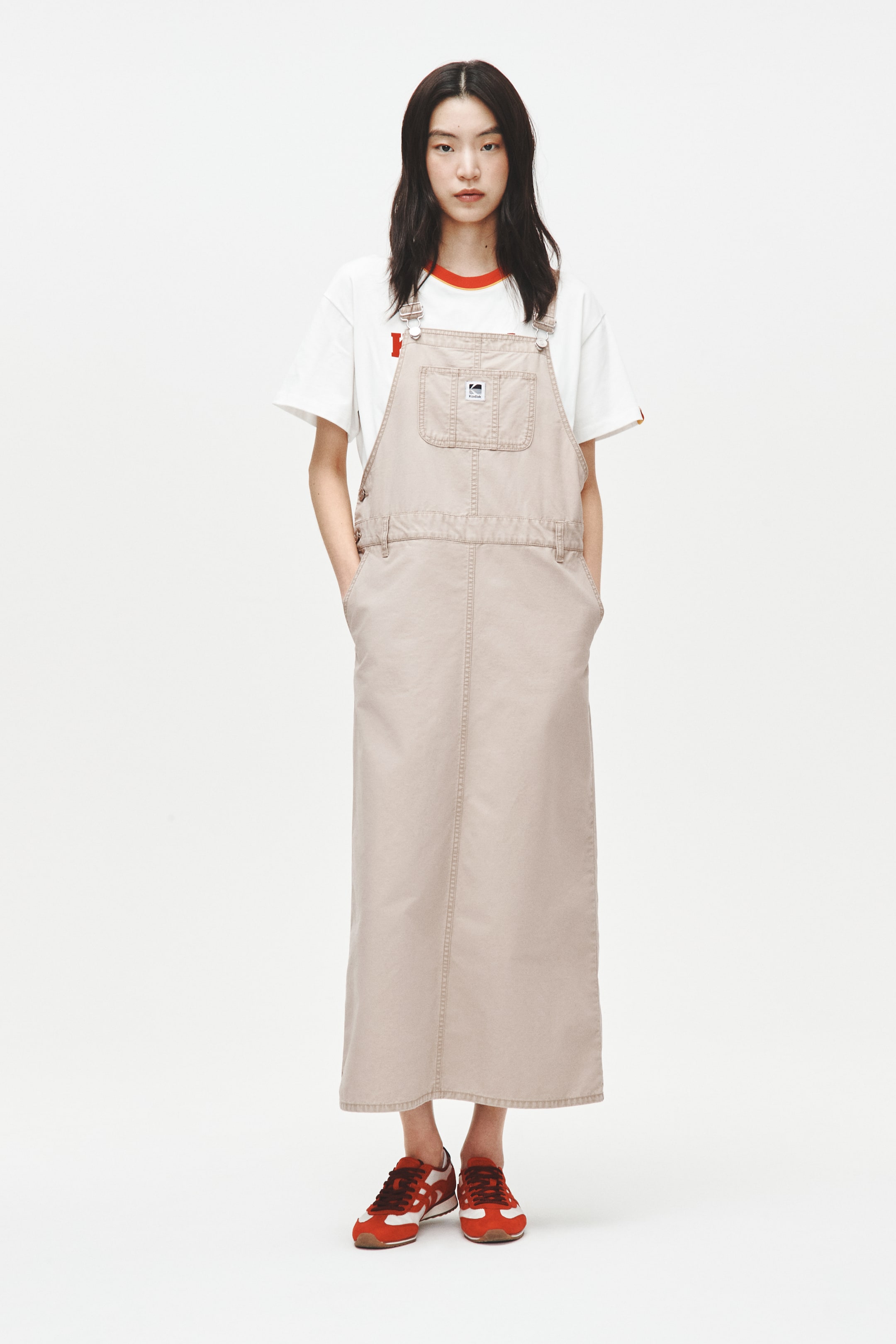 View larger image: A long beige overall dress is showcased from a front angle, featuring adjustable shoulder straps with silver-toned buckles, a large bib pocket with a small rectangular label, and side pockets on the skirt. A white t-shirt with a red-trimmed collar is layered underneath the dress.