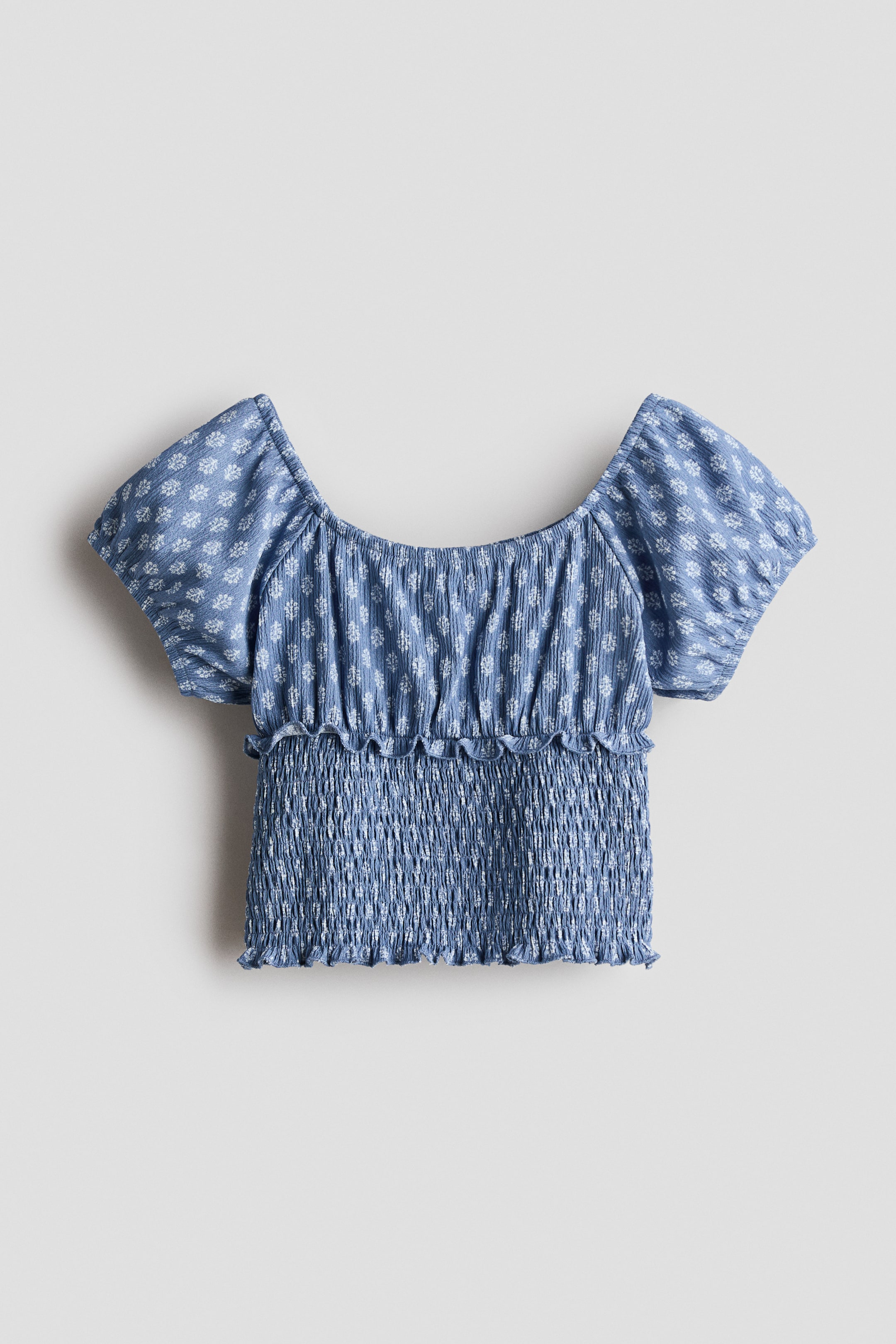 Zobacz większy obraz: Niebieski top typu off-shoulder z krótkimi, bufkowymi rękawami i marszczonym górnym korpusem, ozdobiony białym geometrycznym wzorem kwiatowym. Przód topu prezentuje mocno marszczoną środkową część, również wzorzystą, która prowadzi do falbaniastego dołu.