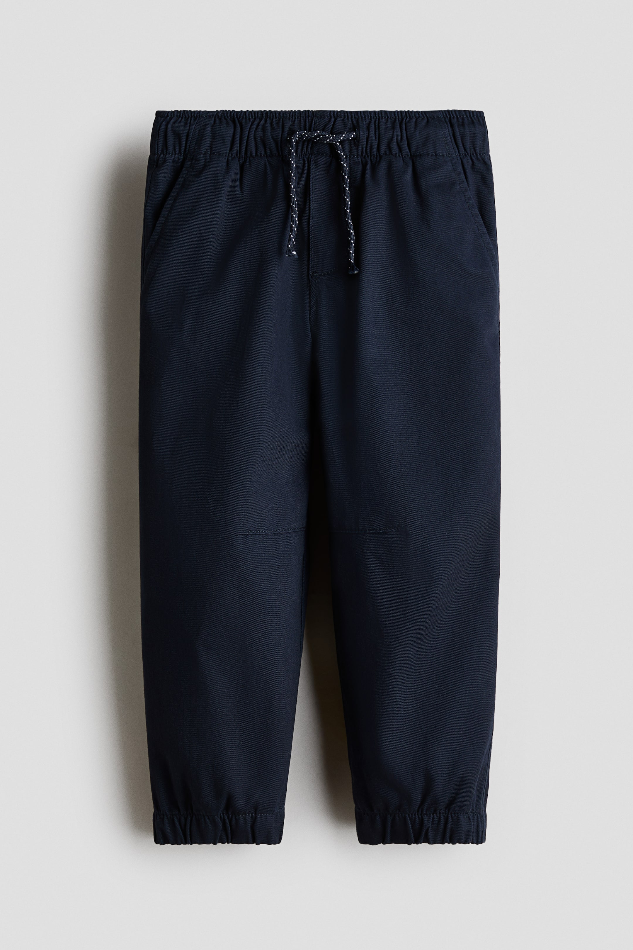 View larger image: Dark blue pants feature an elasticated waistband with a black and white flecked drawstring, two angled front pockets, and stitched knee panels. Elasticated cuffs gather at the ankles.