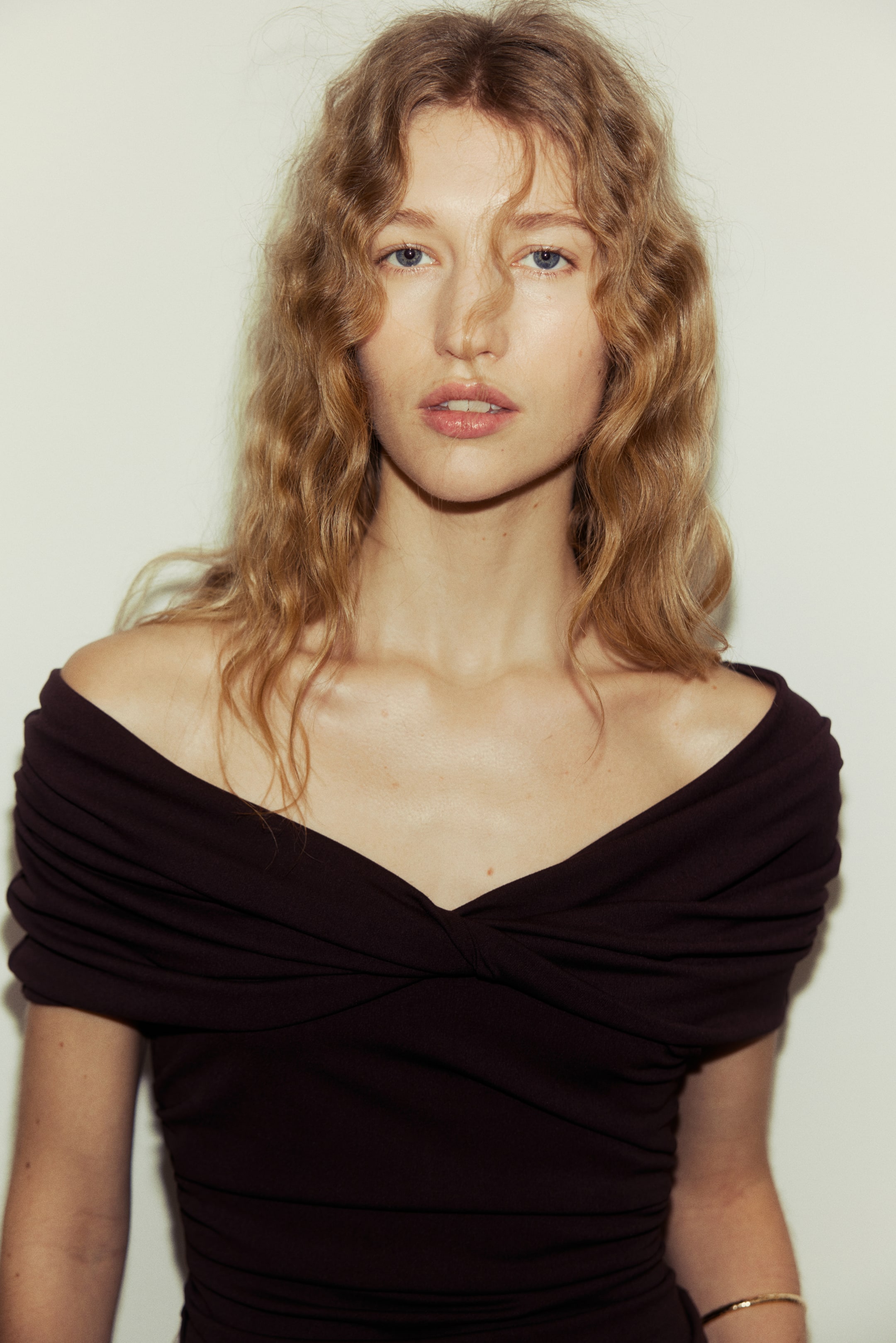 Ver imagen más grande: Un top off-the-shoulder de color marrón oscuro presenta un escote en V retorcido en el centro delantero. La tela se drapea sobre los hombros y se frunce a lo largo del cuerpo, creando una silueta entallada.