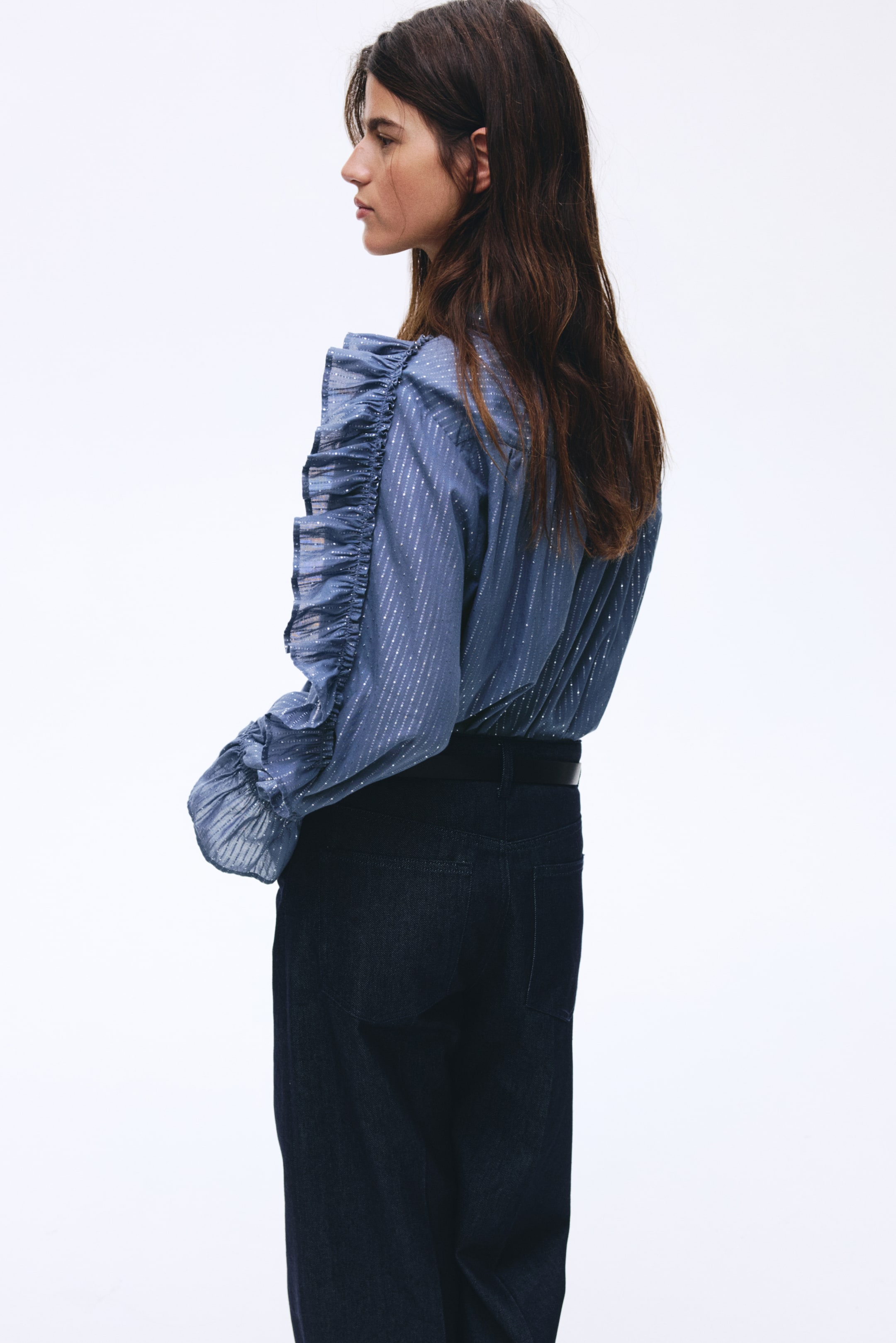 Agrandir l'image: Un haut bleu à manches longues est présenté de côté, avec des rayures diagonales scintillantes et des volants ornant les manches. Il est porté rentré dans un pantalon foncé, maintenu par une ceinture noire.