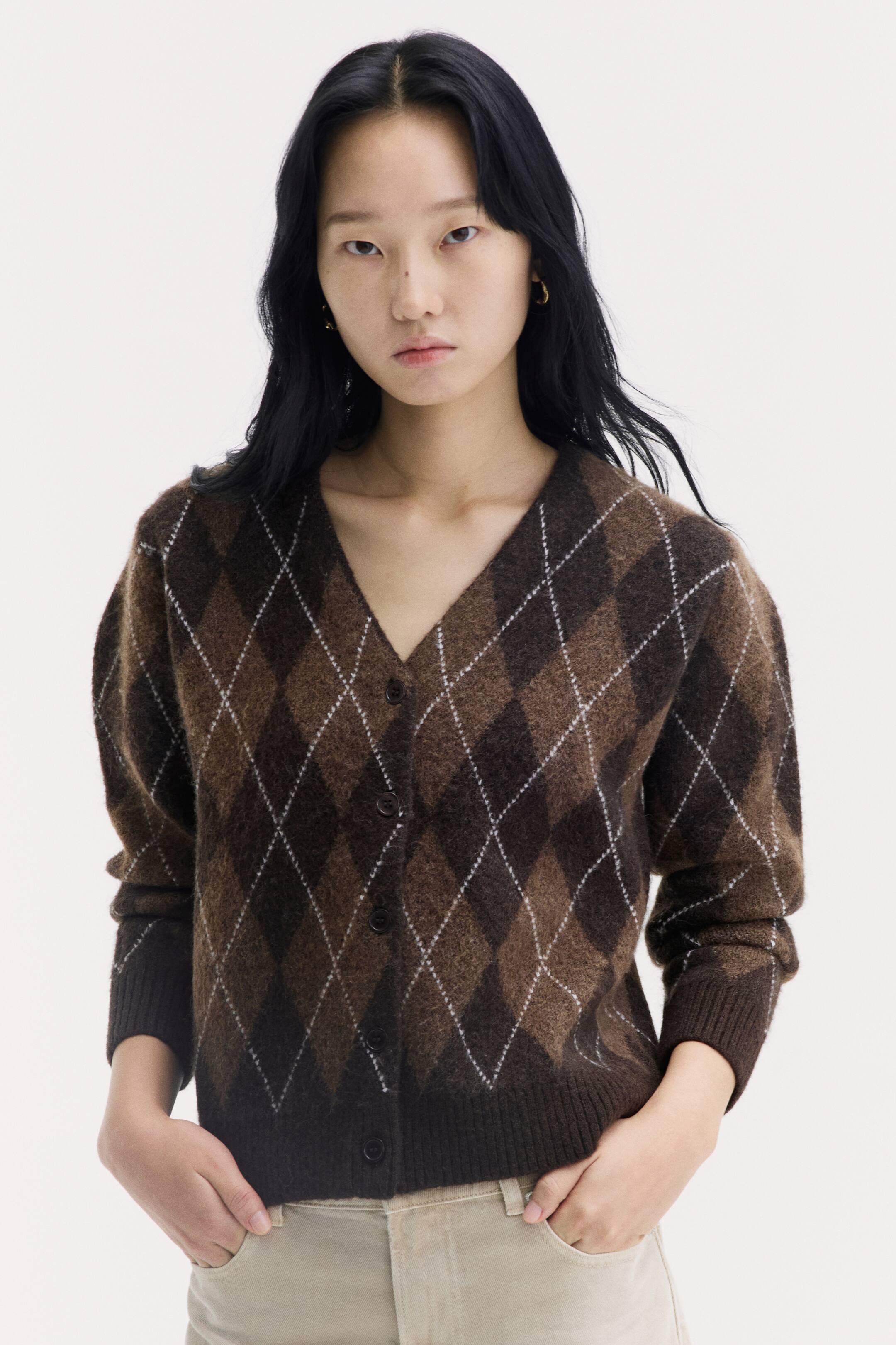 이미지 확대 보기: 모델이 브라운 아가일 패턴의 자카드 니트 카디건을 착용하고 있으며, V넥 라인, 전면 버튼 여밈, 립 처리된 소매단과 밑단이 특징입니다. 모델은 밝은 색상의 트라우저 주머니에 손을 넣은 포즈를 취하고 있습니다.