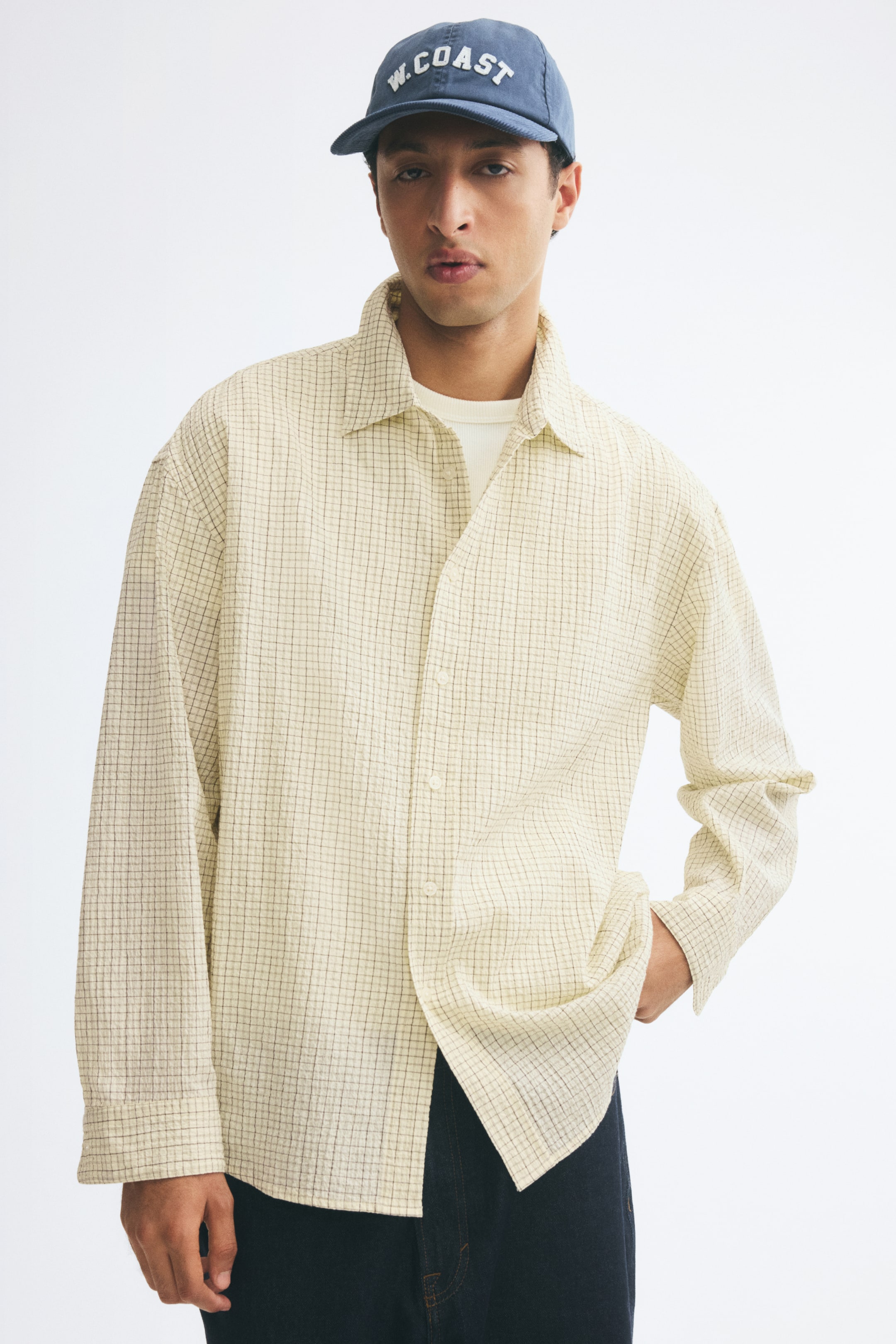 View larger image: A light-coloured shirt with a fine grid pattern is being worn by the model over a white T-shirt. The shirt features a relaxed fit, a turn-down collar, and long sleeves, and is styled with dark trousers and a blue cap.