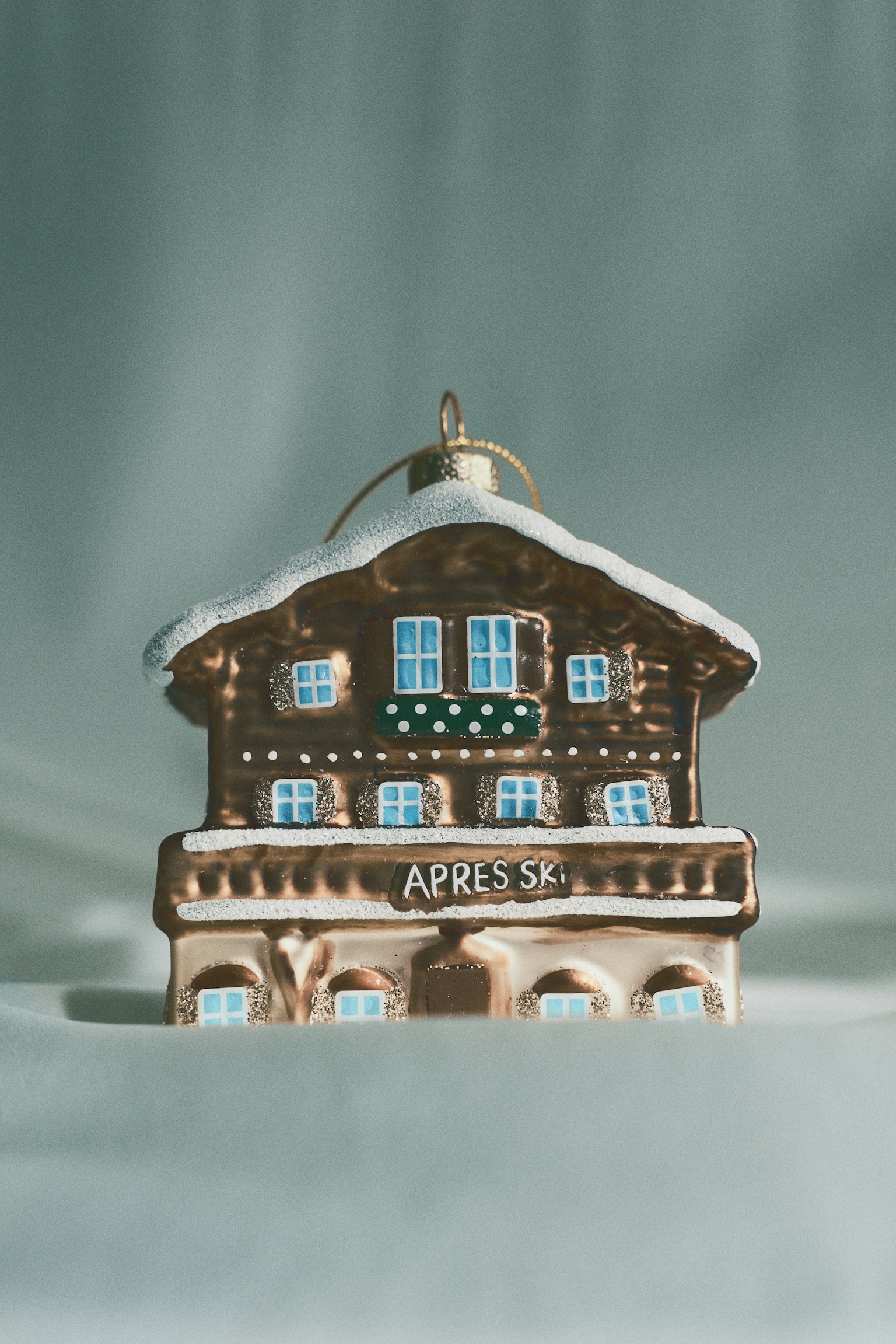 A glass Christmas ornament shaped like a winter chalet is facing forward, showcasing a metallic brown finish with a glittery, snow-covered roof and blue-painted windows. The ornament displays the text "APRES SKI" across the front balcony and features a gold hanging cap and loop at the top. - 1
