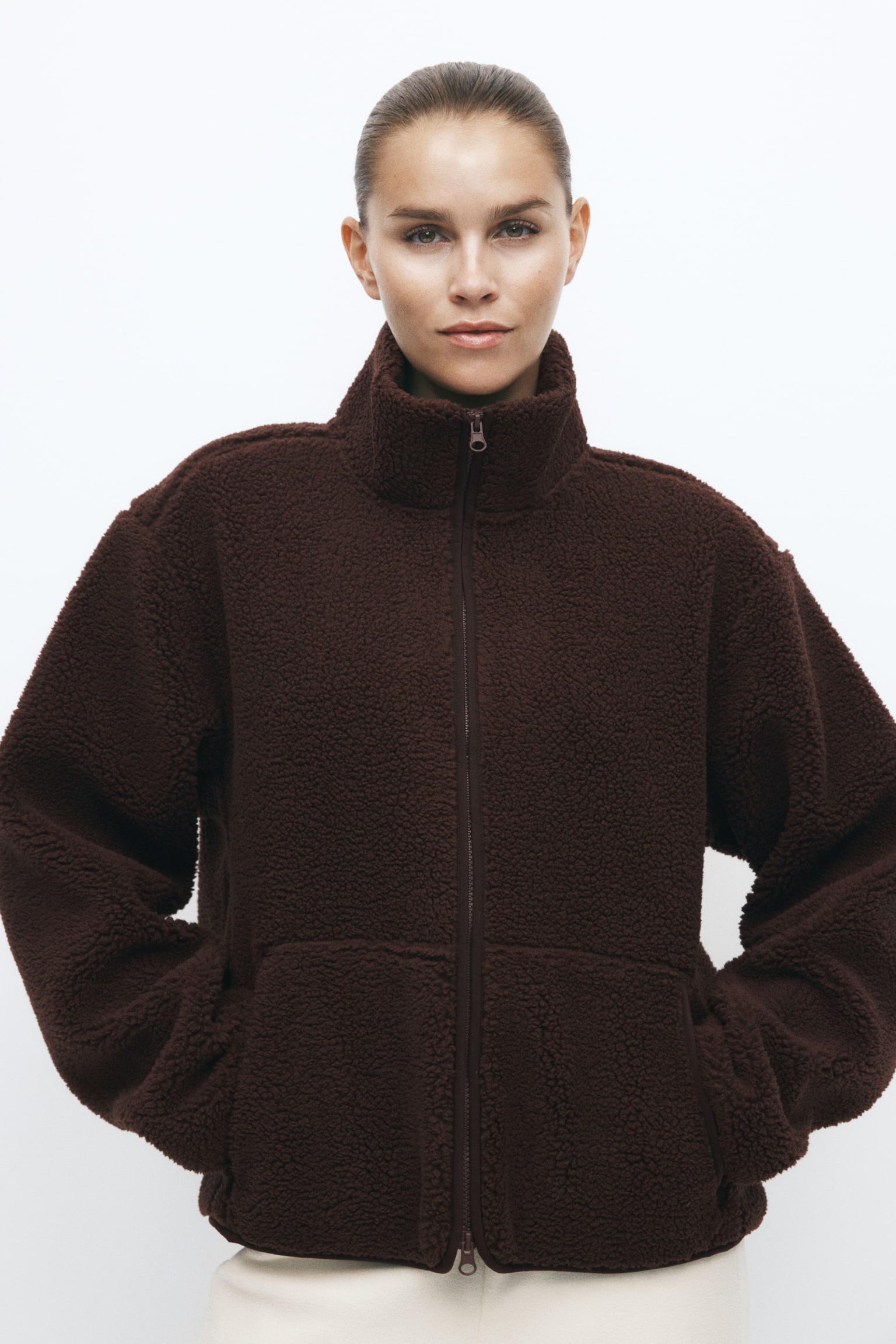 A dark brown, textured fleece jacket with a high collar and front zip is being worn by the model, who has their hands placed in the side pockets. The jacket is styled with cream-coloured trousers. - 1