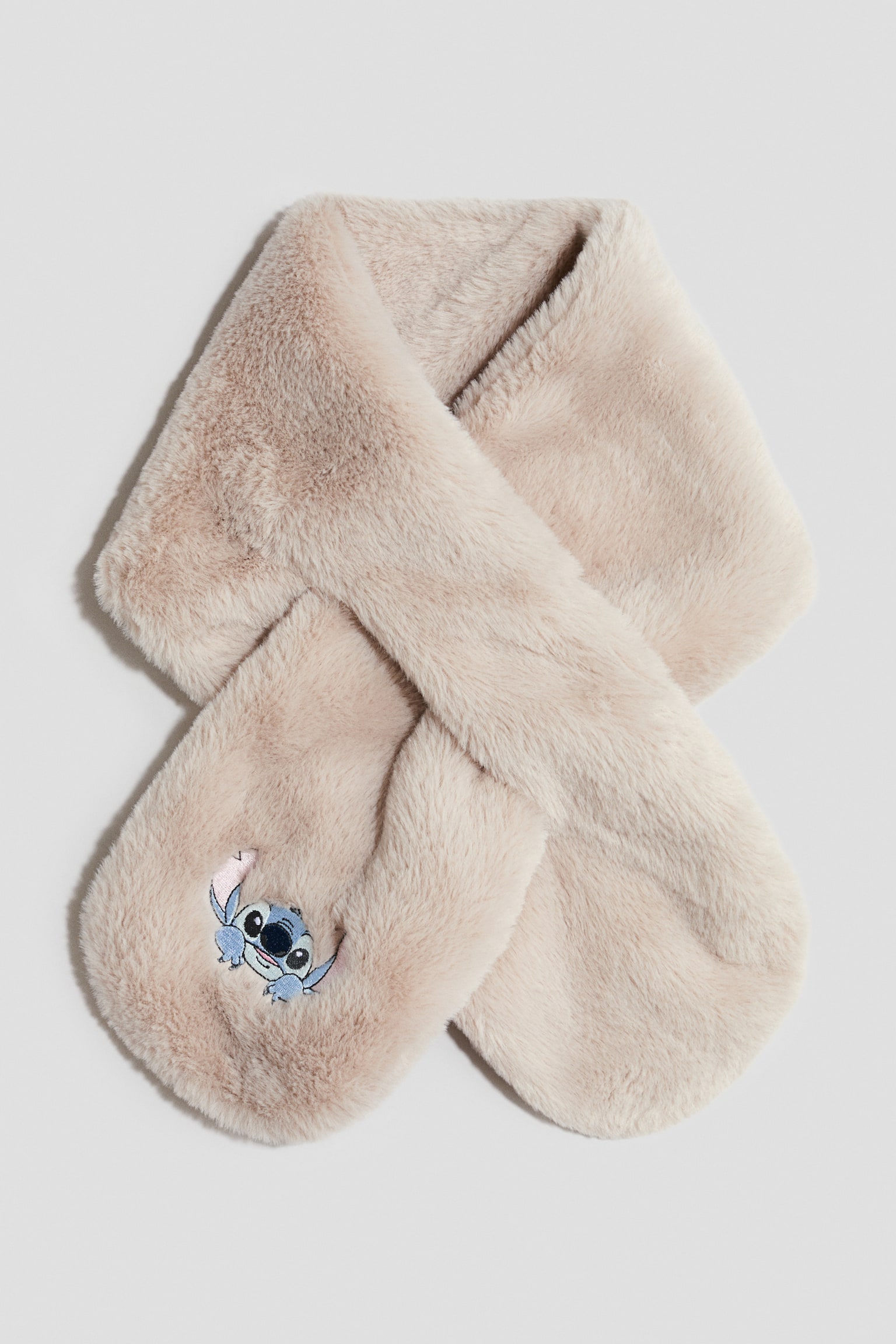 Et lysebeige faux fur-halstÃ¸rklÃ¦de ligger fladt og viser sit forside-design, krydset over sig selv gennem en nÃ¸glehulsÃ¥bning. Det blÃ¸de, fluffy halstÃ¸rklÃ¦de har afrundede ender med en lille blÃ¥ figur broderet nÃ¦r en af spidserne.