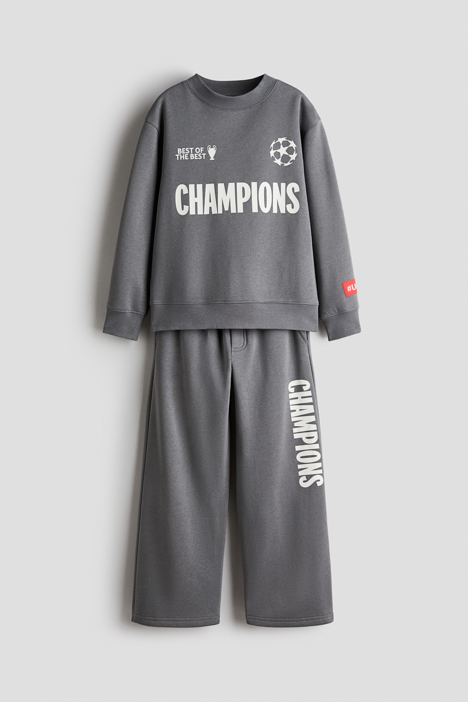 A grey two-piece loungewear set is shown from the front, featuring a long-sleeved crew-neck sweatshirt and matching wide-leg sweatpants. The sweatshirt features a white "CHAMPIONS" graphic across the chest, flanked by a trophy icon and a starball logo, while the sweatpants feature the word "CHAMPIONS" printed vertically in white down the left leg.