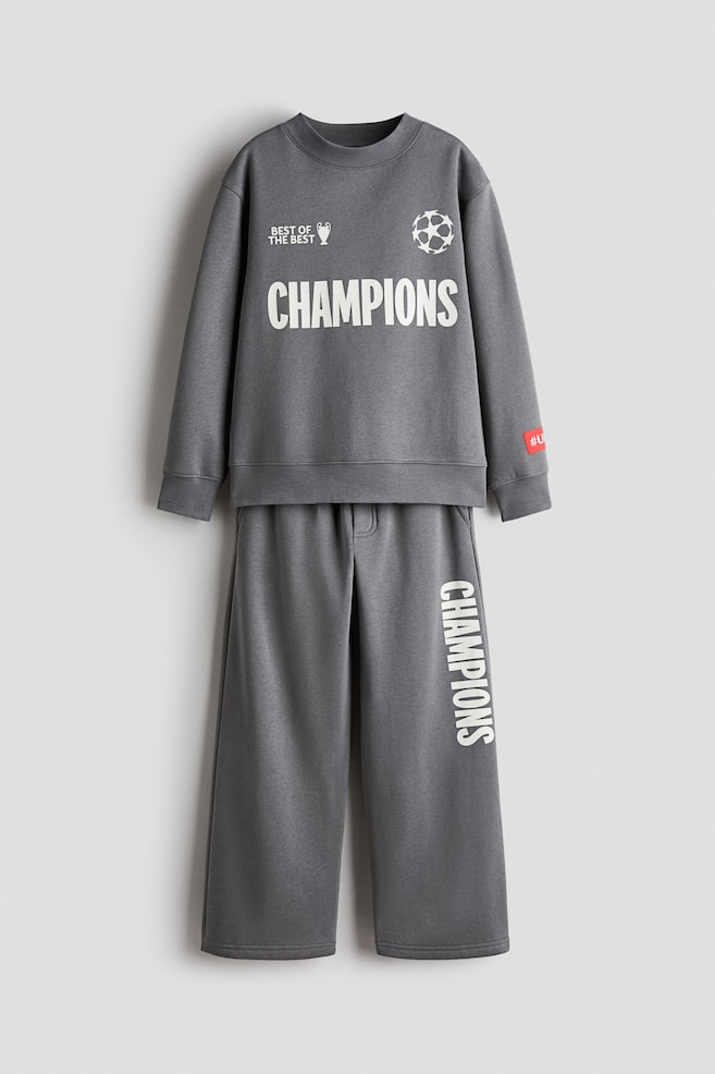 A grey two-piece loungewear set is shown from the front, featuring a long-sleeve crewneck sweatshirt and matching wide-leg sweatpants. The sweatshirt displays a white "CHAMPIONS" graphic across the chest, flanked by a trophy icon and a starball logo, while the sweatpants feature the word "CHAMPIONS" printed vertically in white down the left leg. - 1