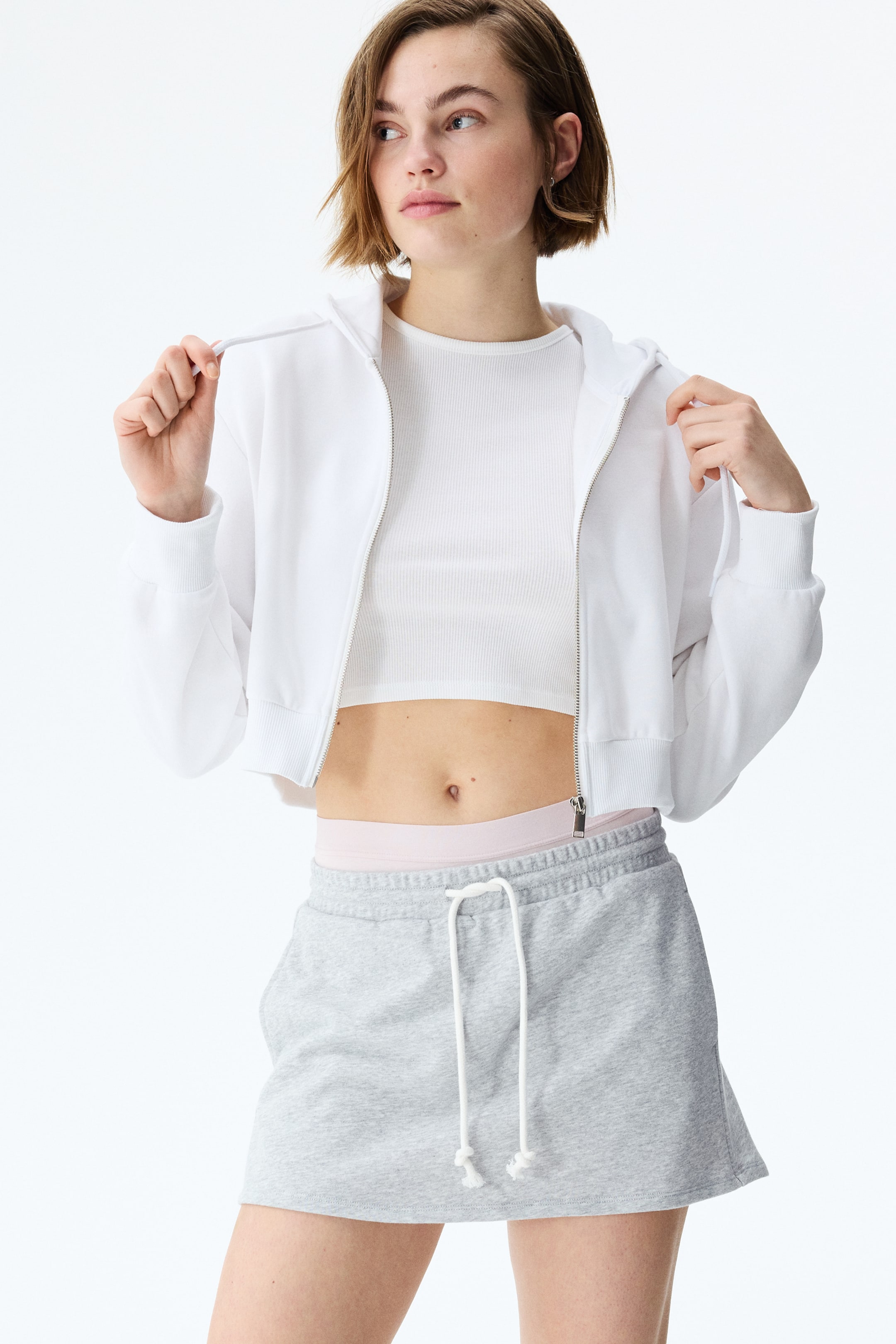Xem ảnh lớn hơn: Một chiếc áo hoodie trắng dáng lửng có khóa kéo phía trước mở một phần, cổ tay áo và gấu áo có gân, được mặc bên ngoài áo crop top cổ tròn trắng có gân. Một chiếc váy mini chất liệu nỉ xám có cạp chun với dây rút trắng.