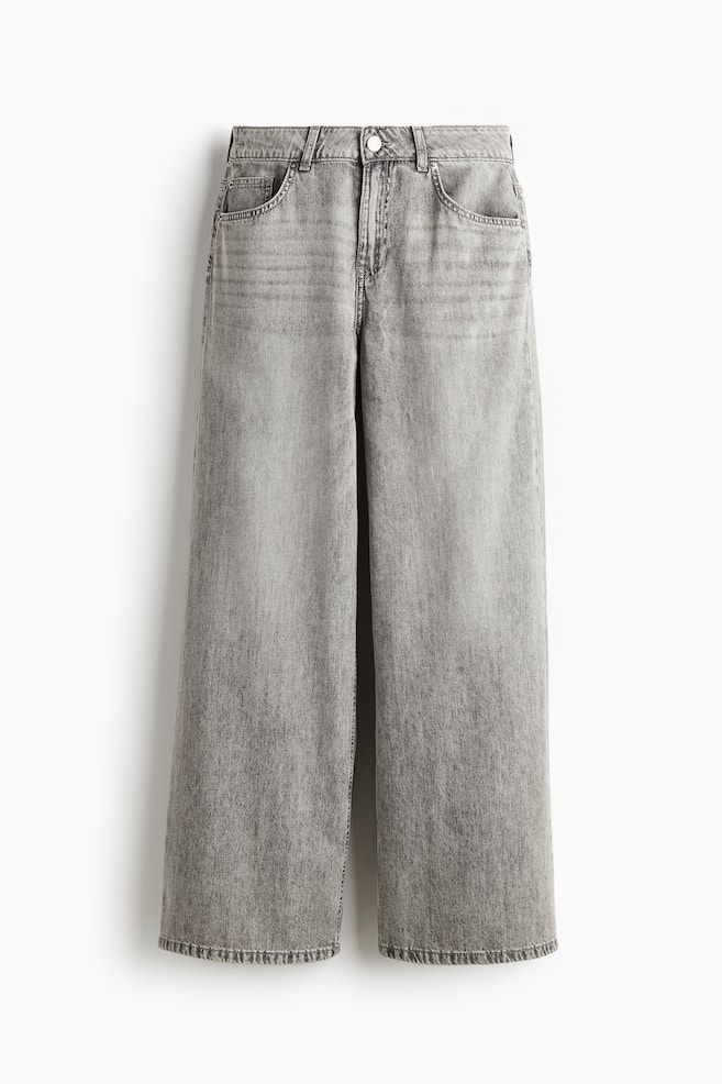 Önden görünümde gri 5 cepli bir kot pantolon çifti. Yapısal bir denimden üretilmiş olup, düğme ve fermuar kapamalı normal bir bele, iki ön cebe ve bir bozuk para cebine sahiptir. Kot pantolonlar, belden kalçaya kadar normal görünüp, uyluktan eteğe kadar genişleyen geniş paça kesimi ile uzun bir uzunlukta tasarlanmıştır. - 1