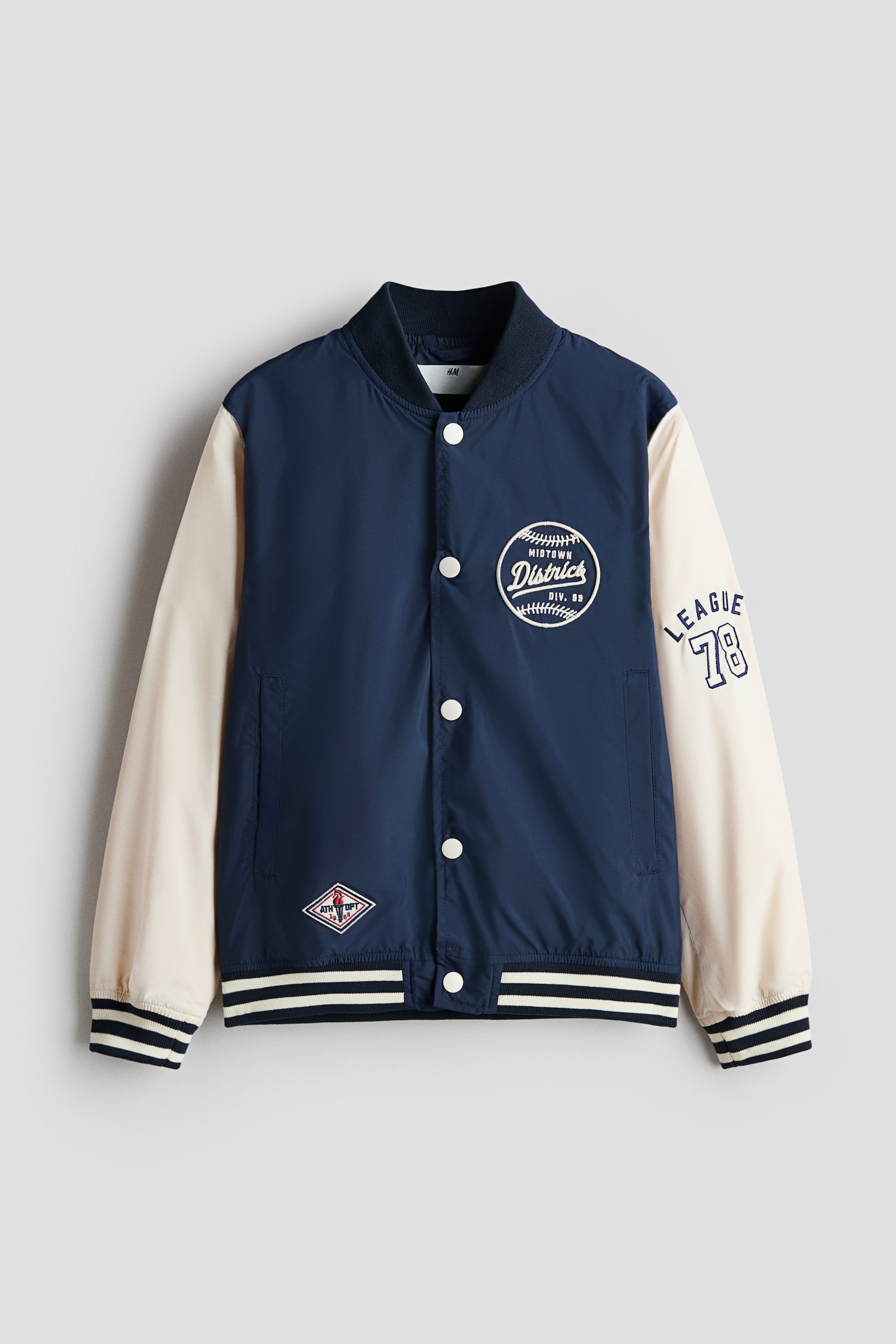 Visualizza immagine più grande: La giacca varsity è mostrata frontalmente, con il corpo blu navy e maniche crema a contrasto, un colletto a costine blu navy e chiusure con bottoni a pressione bianchi. Presenta polsini e orlo a costine con righe blu navy e crema, una toppa circolare "Midtown District" sul petto sinistro e "League 78" ricamato sulla manica destra.