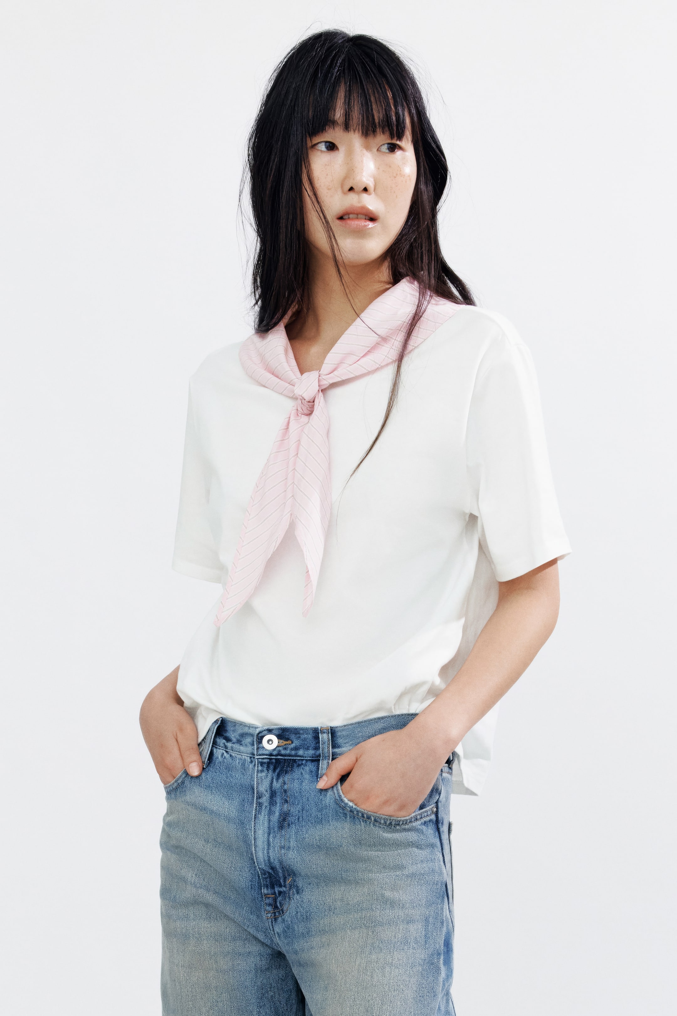 View larger image: The model is wearing a white T-shirt made from soft cotton jersey, featuring a round neckline, subtly dropped shoulders, and a straight-cut hem. A pink striped woven scarf, attached to the collar, is elegantly tied around the model's neck. The T-shirt is partially tucked into light wash jeans.