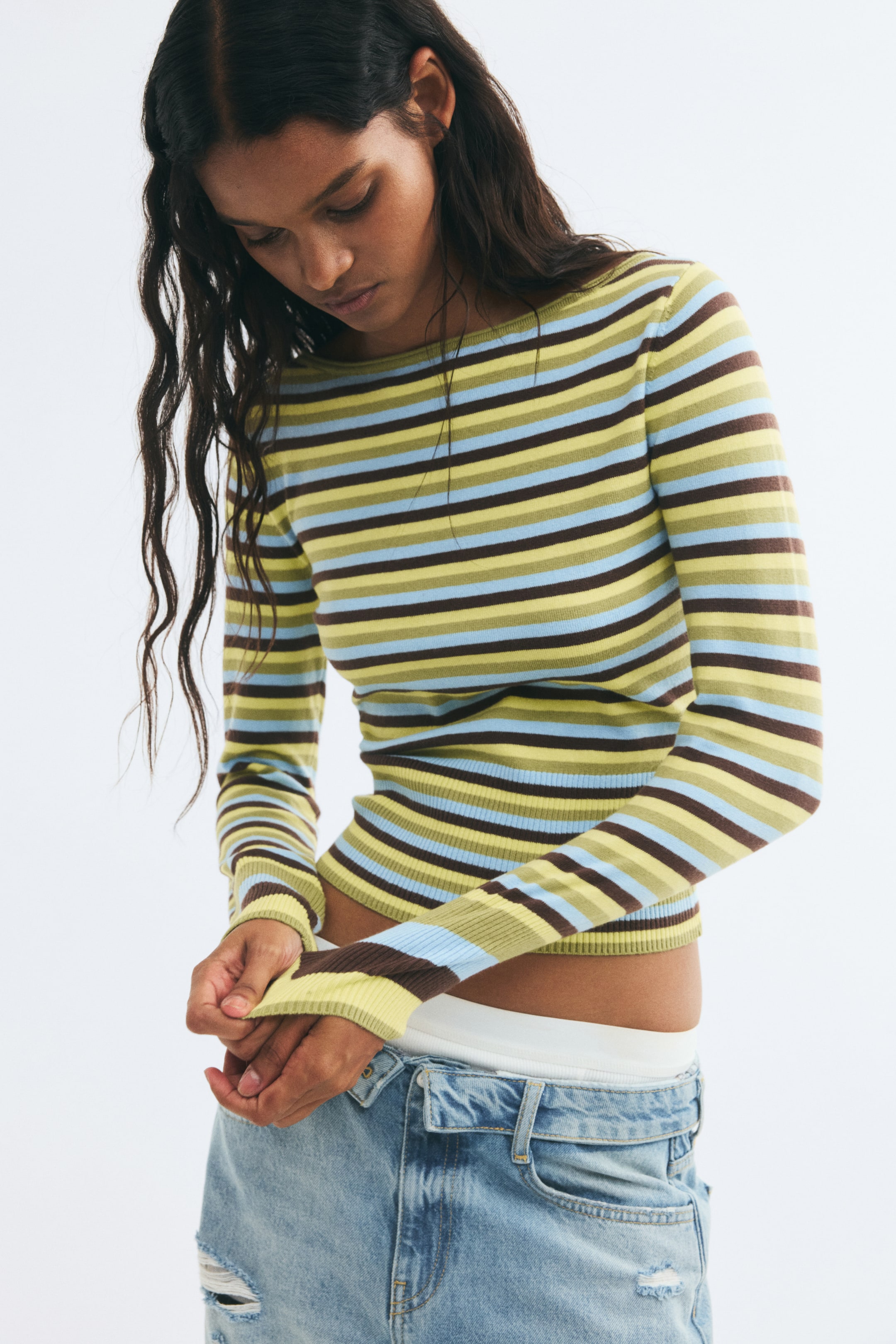 Voir une image plus grande: Un haut en tricot ajusté à manches longues avec des rayures horizontales vertes, jaunes, bleues et brunes est porté par le mannequin, qui ajuste le poignet côtelé d’une manche. Le haut court présente un col bateau et est agencé avec un jean en denim délavé clair.