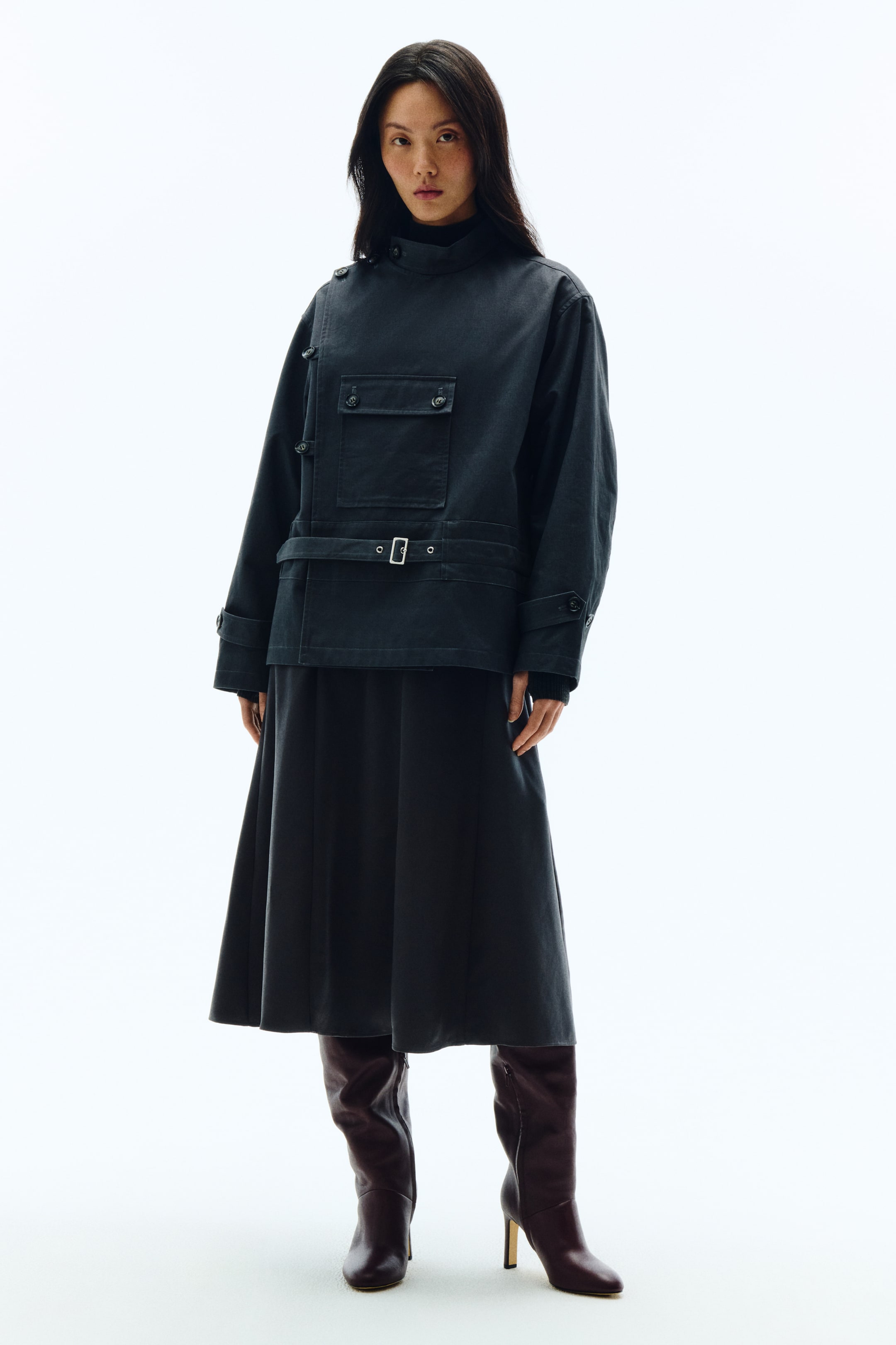拡大画像を表示: ダークカラーのジャケットに、ハイスタンドカラー、ショルダーボタンのディテール、フラップ付きチェストポケット、シルバーバックル付きベルトウエストを備えたフルコーディネート。ブラックのタートルネックの上にレイヤードし、ダークカラーのプリーツミディスカートとダークブラウンのニーハイヒールブーツを合わせ、すべて正面向きでスタイリングされています。