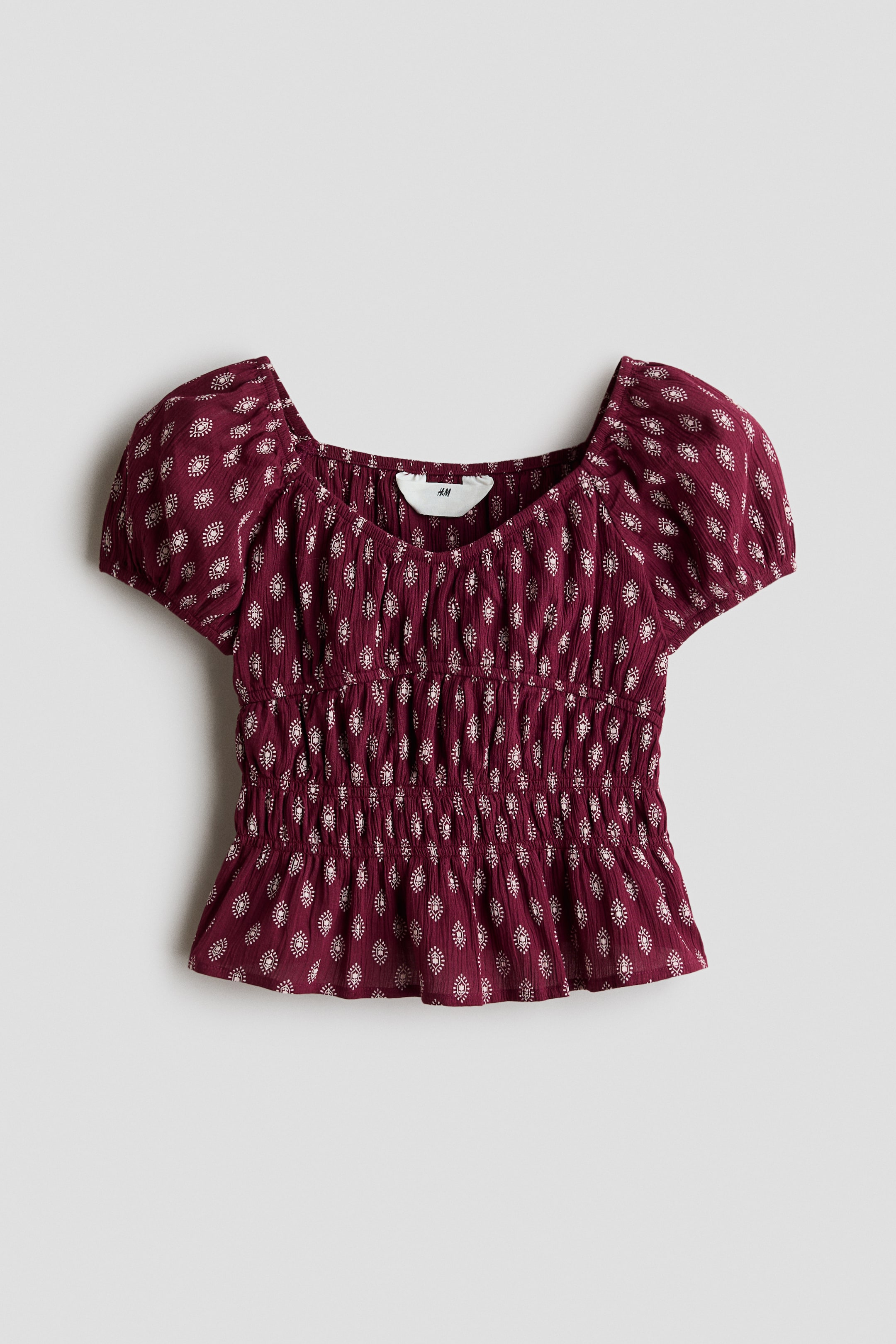 View larger image: A burgundy top features a repeating pattern of small white oval motifs. It showcases a square neckline, short puffed sleeves, and a shirred bodice that creates a fitted, textured appearance.