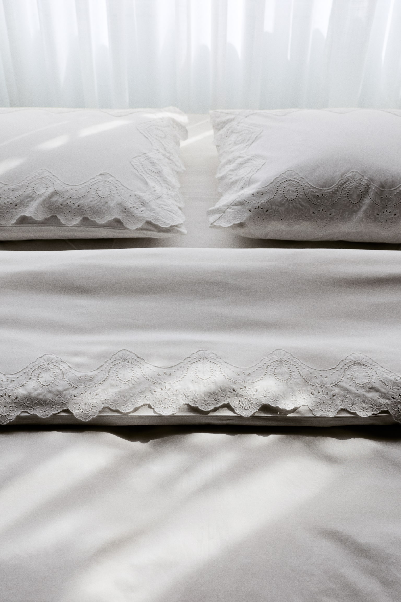 "Displayed is a white bedding set, highlighting a pillowcase adorned with a scalloped eyelet lace border on top. Beneath it, a folded white sheet or duvet cover showcases a ruffled scalloped edge with eyelet lace, layered over another folded white sheet with a scalloped edge." - 1