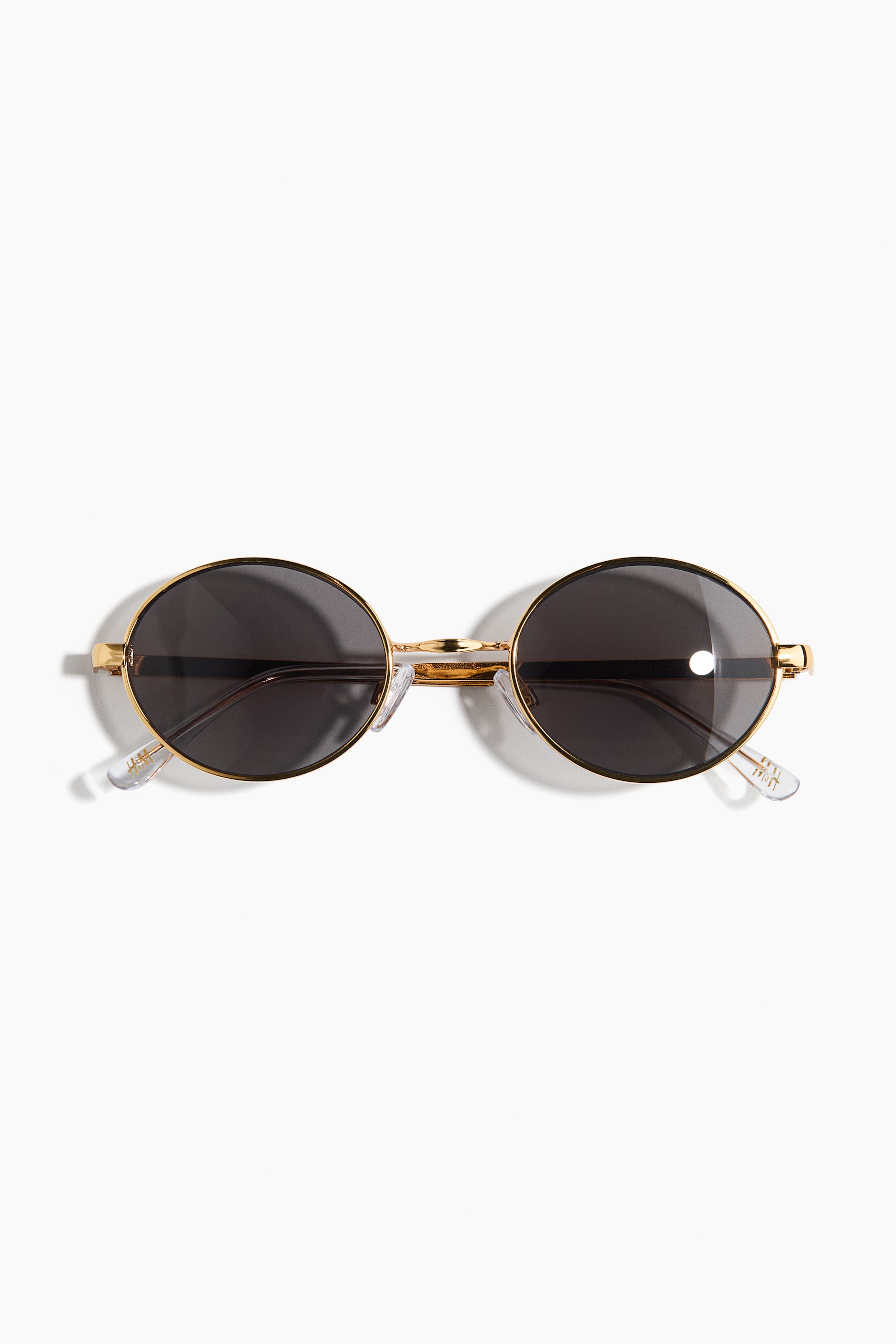 View larger image: Oval sunglasses featuring slim gold-toned metal frames and dark-tinted, UV-protective plastic lenses. They are presented with adjustable clear nose pads and metal sidepieces ending in clear plastic tips, facing FRONT.