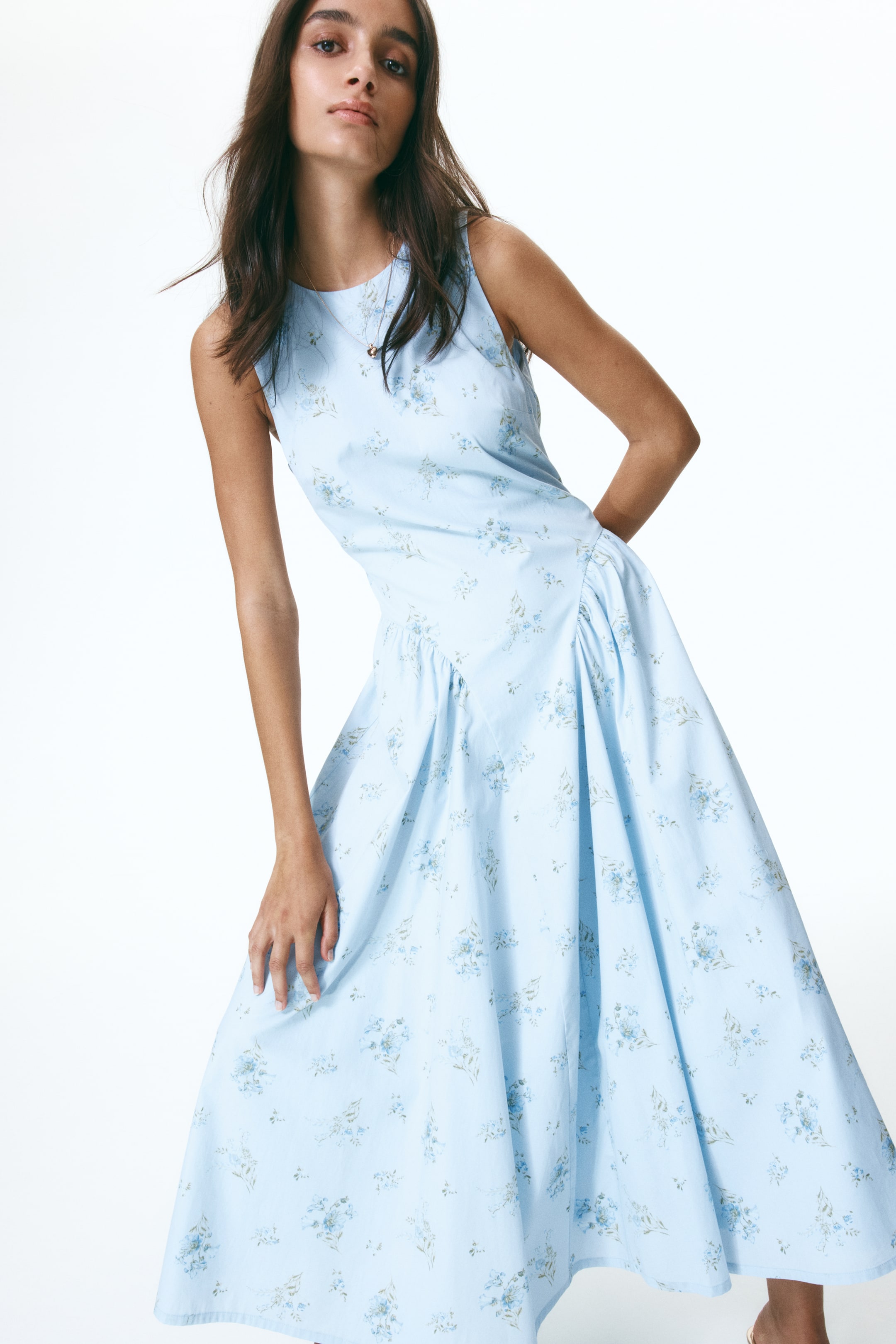 View larger image: The model is wearing a calf-length, sleeveless dress made from light blue cotton poplin, adorned with a floral print. This dress showcases a round neckline, a fitted bodice, and a flared skirt with gathers over the hips. The model's hand rests gracefully on the skirt, enhancing its elegant flow.