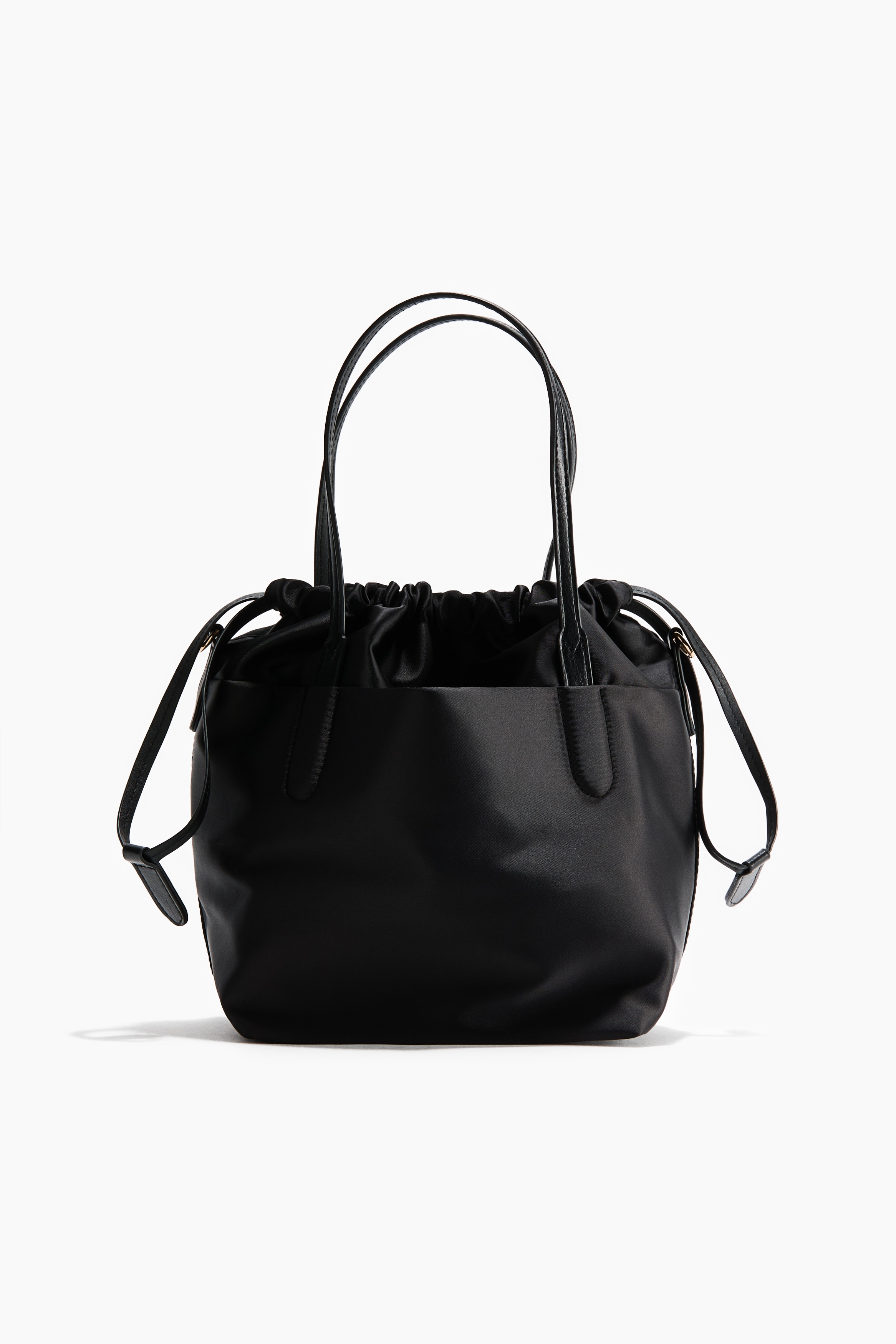 A black bucket bag with a drawstring top and long, narrow handles is being held by the model against a dark, pinstriped blazer. The bag features a soft, slouchy structure with thin straps gathered in the hand and a detachable shoulder strap with a gold-tone carabiner hook.