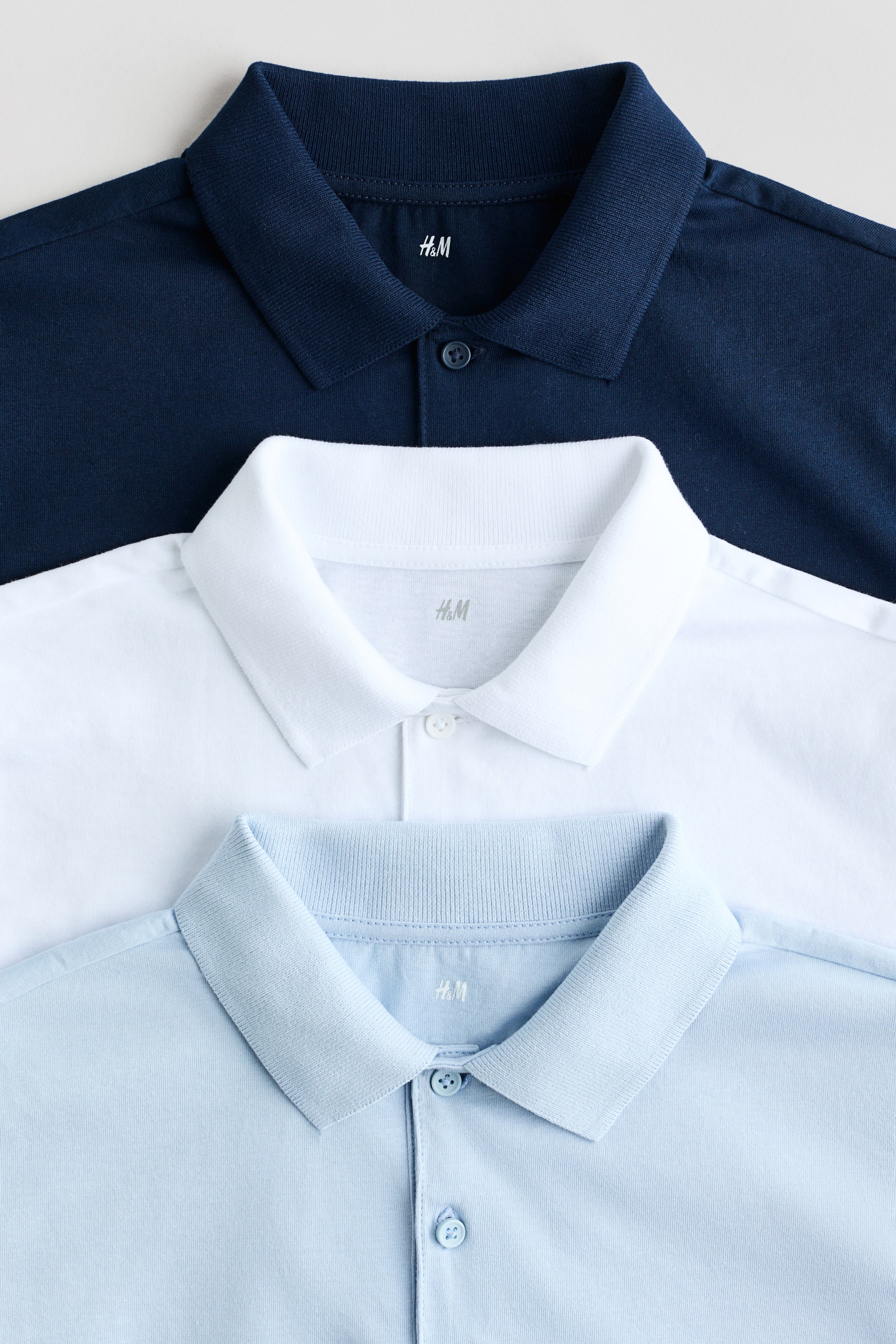 Agrandir l'image: Des polos en jersey de coton doux sont présentés de face, dotés de cols côtelés, de pattes de boutonnage et de manches courtes. Trois polos bleu marine, blanc et bleu clair apparaissent superposés, le haut du buste et le col de chacun étant visibles.