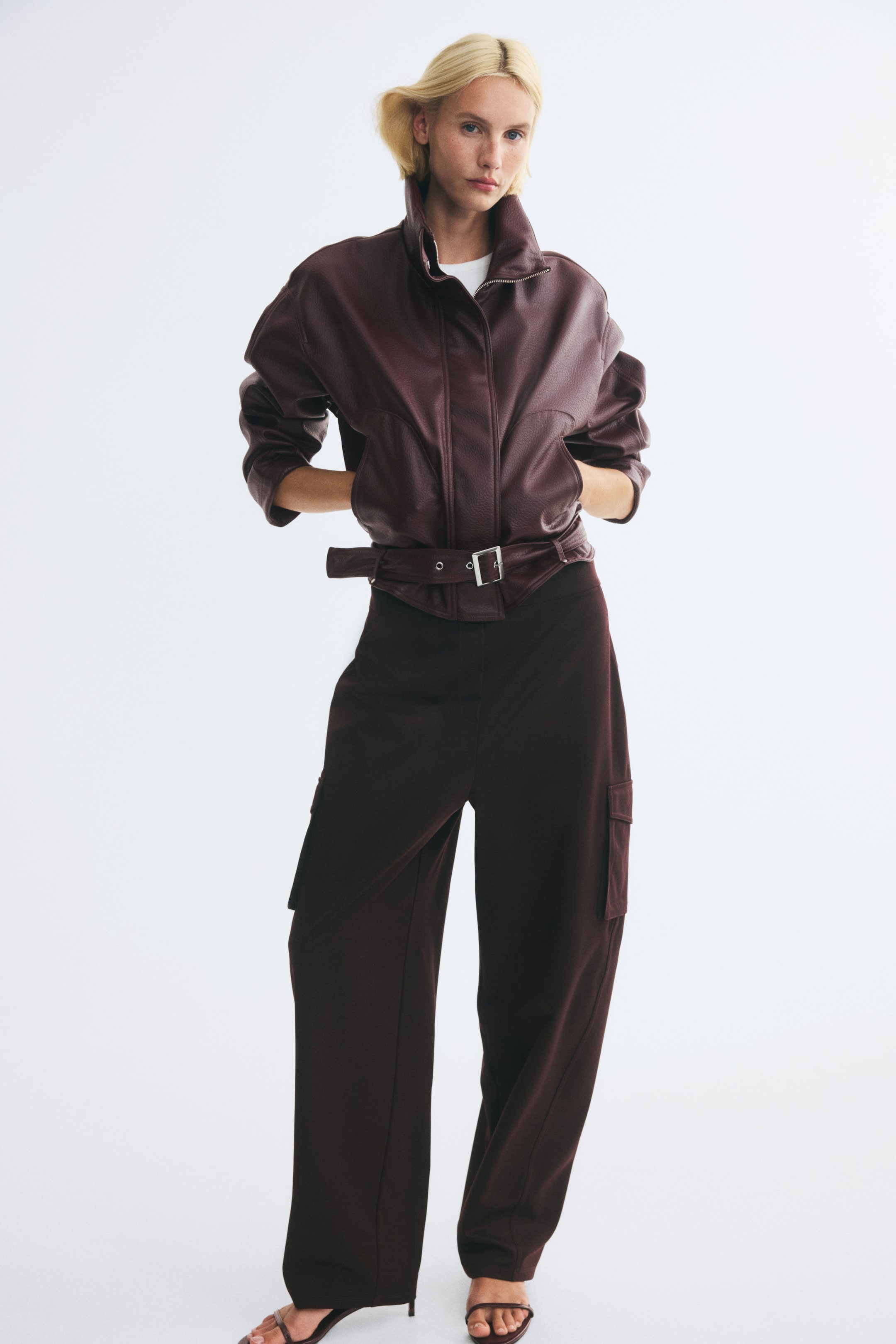 Agrandir l'image: Une veste bordeaux foncé en tissu enduit, dotée d’un col montant, de manches tombantes et d’une ceinture ajustable avec boucle métallique à l’ourlet, est portée par le mannequin. La veste est superposée sur un t-shirt blanc et un pantalon large brun foncé, tandis que les mains du mannequin reposent dans les poches avant.