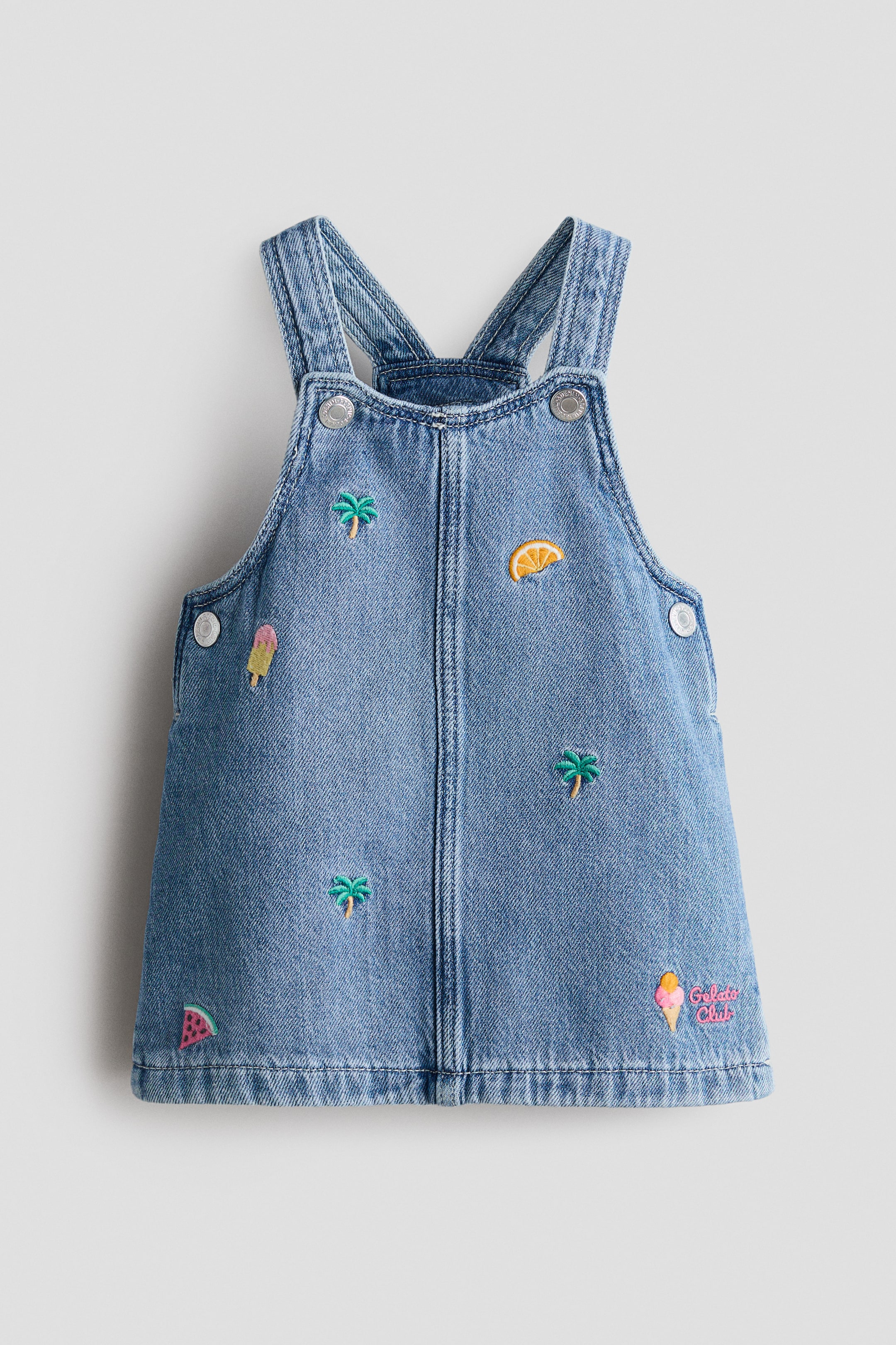 Größeres Bild anzeigen: Ein hellblaues Jeanskleid in Latzhosenstil ist von vorne zu sehen, ausgestattet mit verstellbaren Schulterträgern mit silbernen Knöpfen und seitlichen Knopfverschlüssen. Das Kleid ist mit verschiedenen gestickten Motiven verziert, darunter grüne Palmen, eine orangefarbene Zitrusscheibe, ein pink-gelbes Eis, eine rote und grüne Wassermelonenscheibe sowie der Schriftzug "Gelato Club".