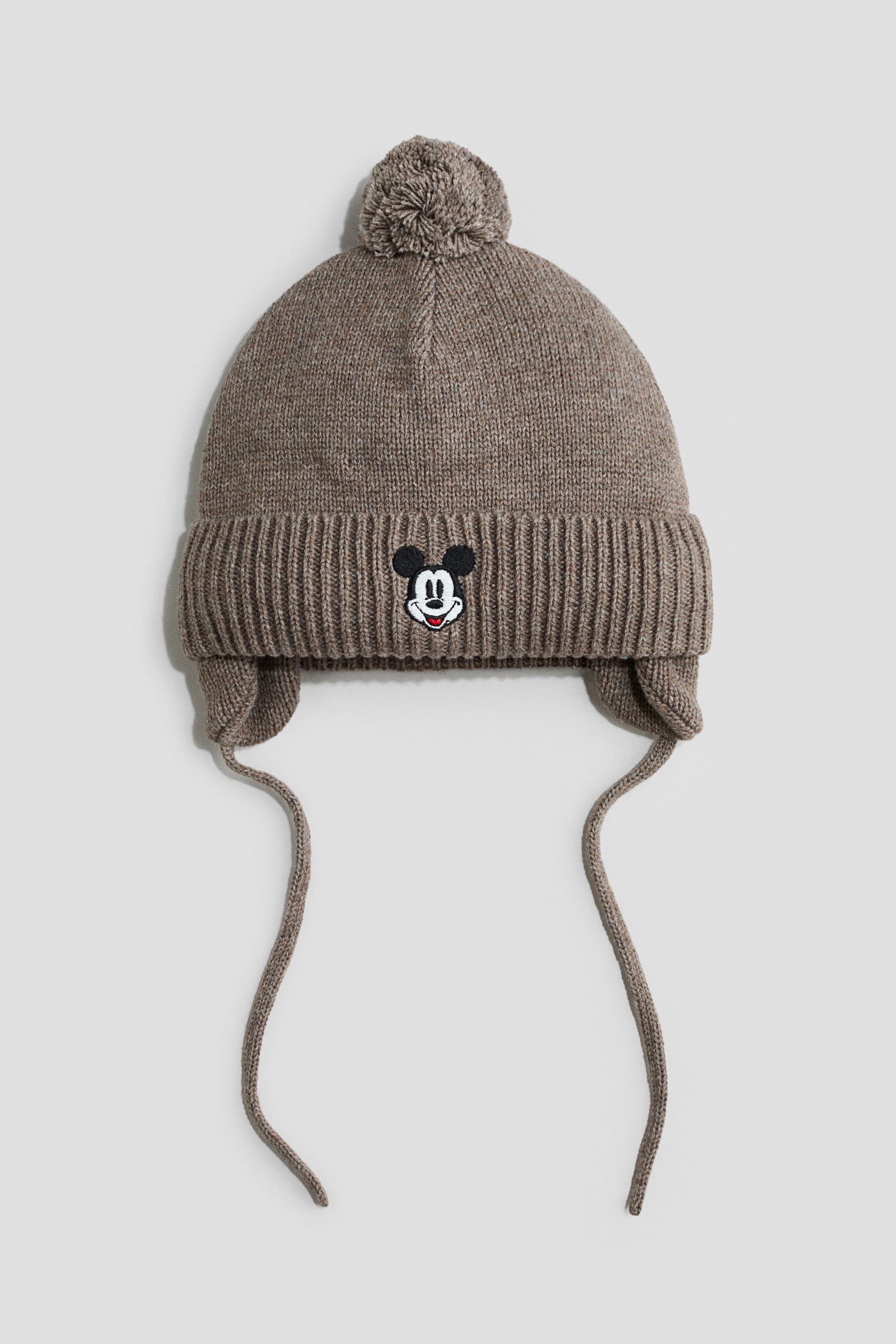 A heather brown knit earflap hat is facing front, featuring a matching pom-pom on top and a wide ribbed fold-over cuff. The center of the cuff displays a small embroidered Mickey Mouse face, with knit earflaps extending down into long hanging ties on both sides.