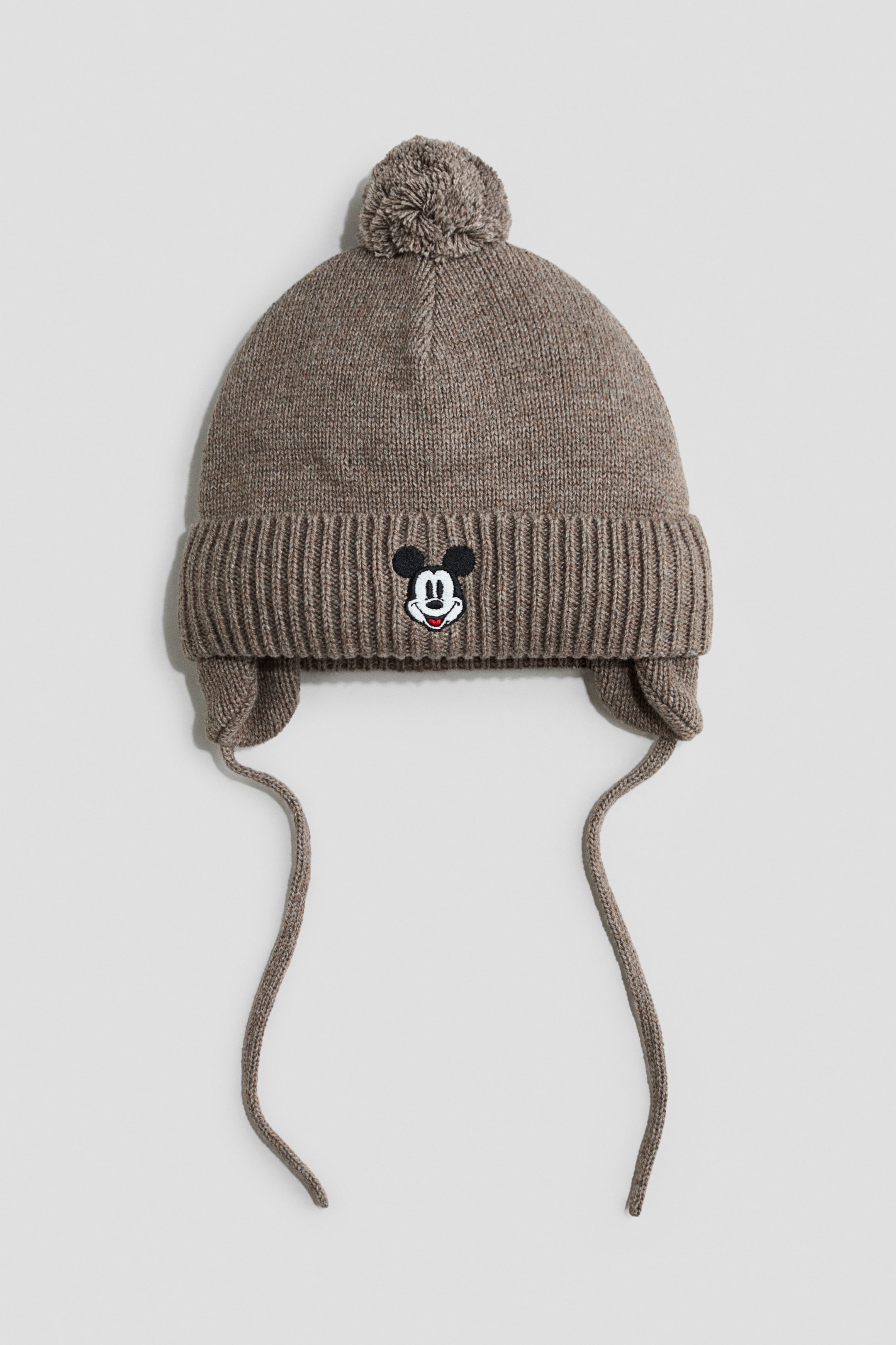 View larger image: A heather brown knit earflap hat is facing front, featuring a matching pom-pom on top and a wide ribbed fold-over cuff. The centre of the cuff displays a small embroidered Mickey Mouse face, with knit earflaps extending down into long hanging ties on both sides.