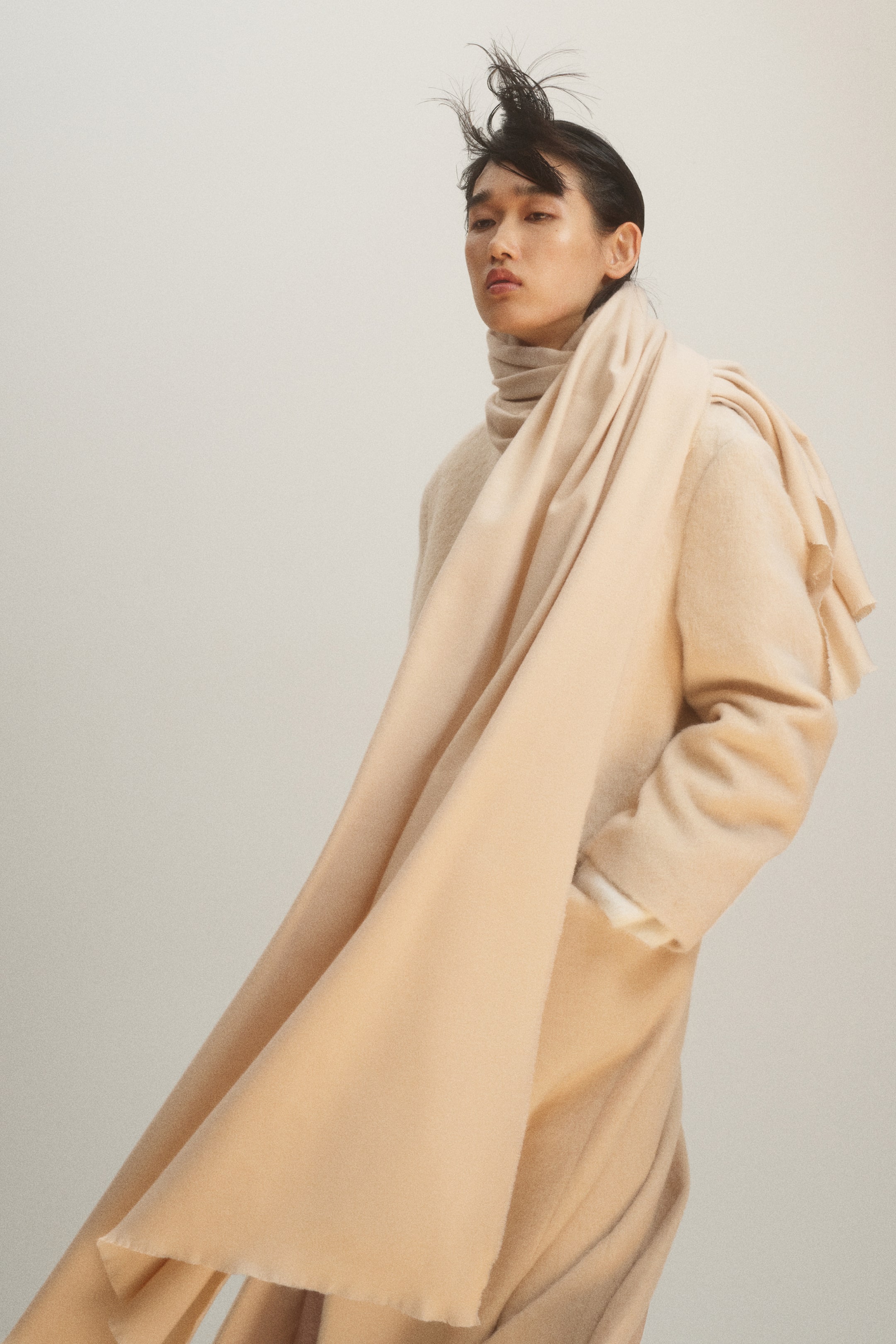 View larger image: A beige blanket scarf in a soft weave with frayed edges is being worn draped over the shoulder and wrapped around the neck of the model. The scarf is being styled as a layer over a matching beige coat.