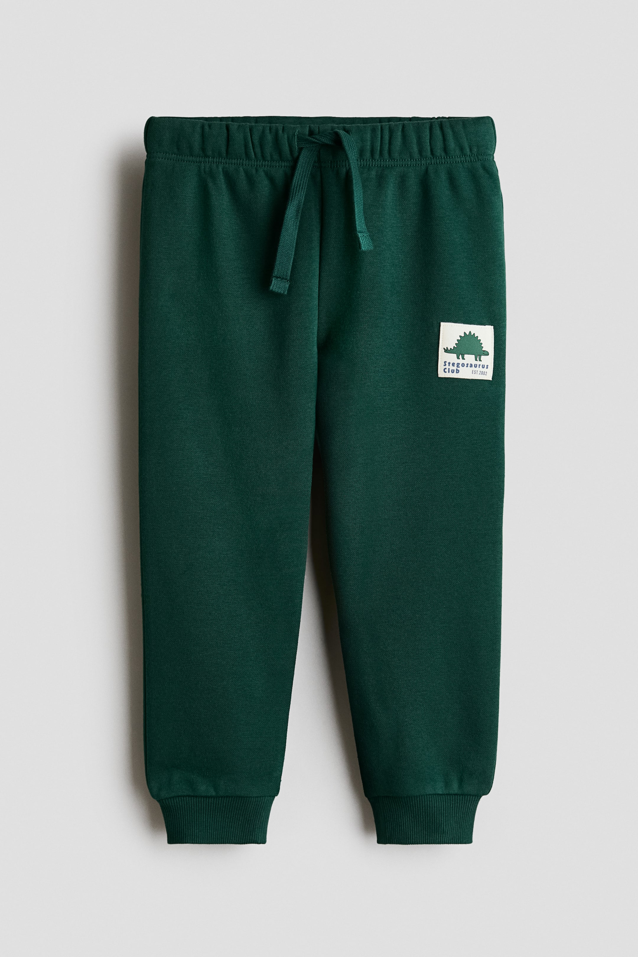 Grotere afbeelding bekijken: De donkergroene joggingbroek wordt van voren getoond en heeft een elastische tailleband met een vastgeknoopte trekkoord. Een wit vierkant embleem met daarop een donkergroene stegosaurusafbeelding en de tekst 'Stegosaurus Club' is zichtbaar op het bovenbeen aan de linkerkant, en de broekspijpen eindigen met geribbelde boorden.