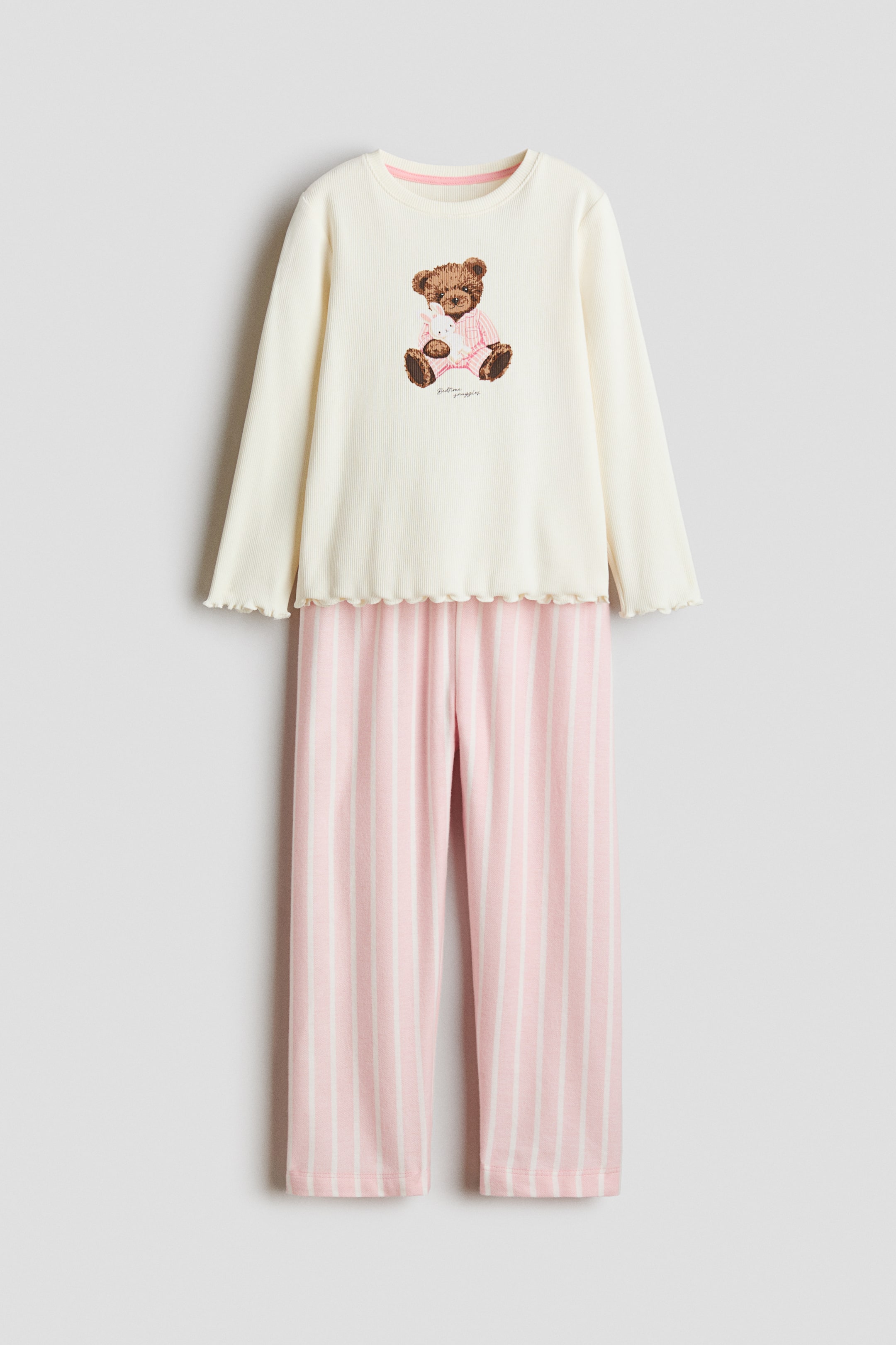 View larger image: Pyjamas, shown from the front, feature an off-white, long-sleeved ribbed top with a round neckline, displaying a teddy bear print on the front, and scalloped edges at the cuffs and hem. The bottoms feature pink and white vertical stripes on straight legs.