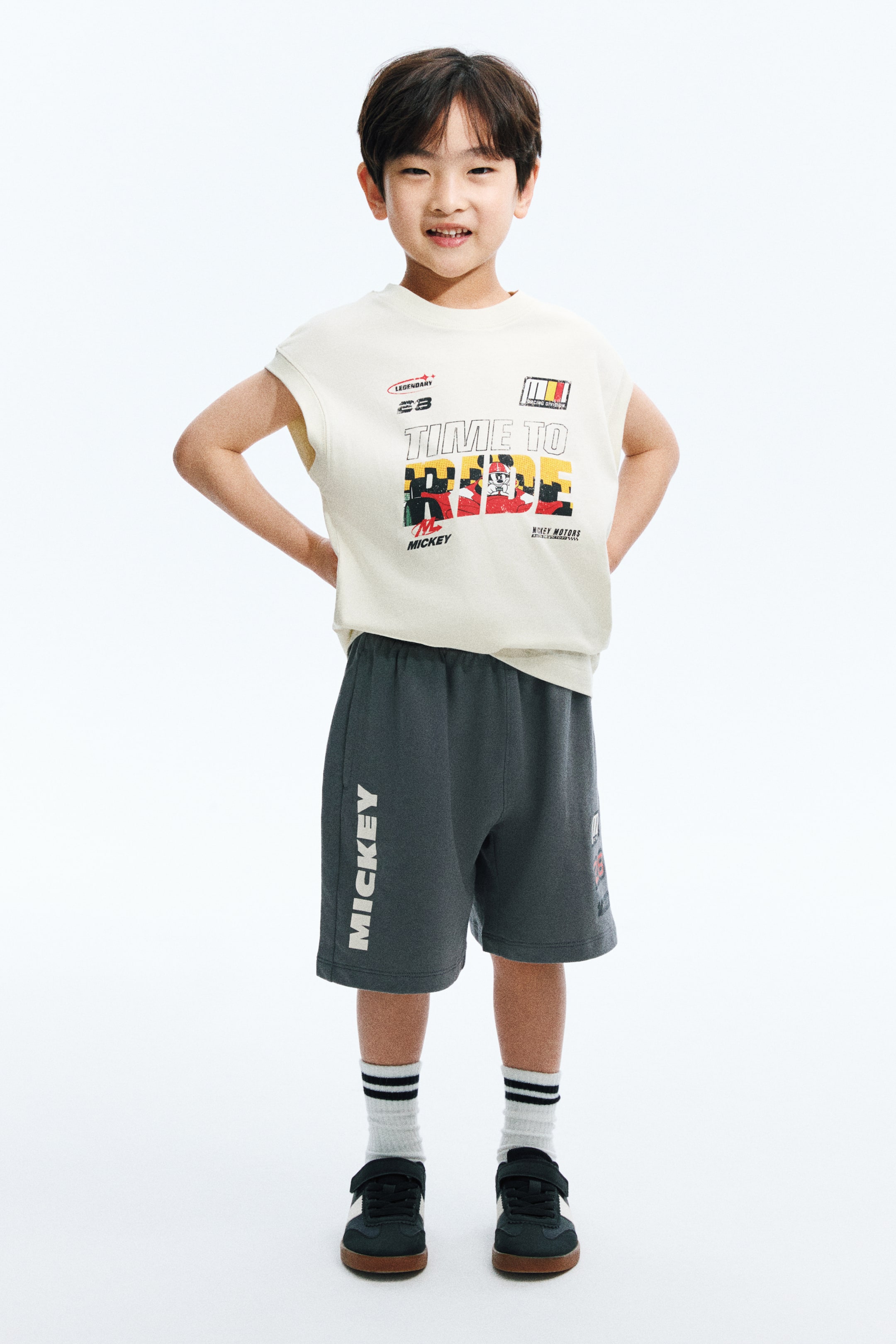 View larger image: A cream sleeveless top paired with dark grey shorts creates a stylish two-piece set. The sleeveless top showcases a crew neck and a front graphic print featuring "TIME TO RACE" alongside a Mickey Mouse motif and racing-themed text. The dark grey shorts come with an elasticated waistband and "MICKEY" printed vertically along the left leg.