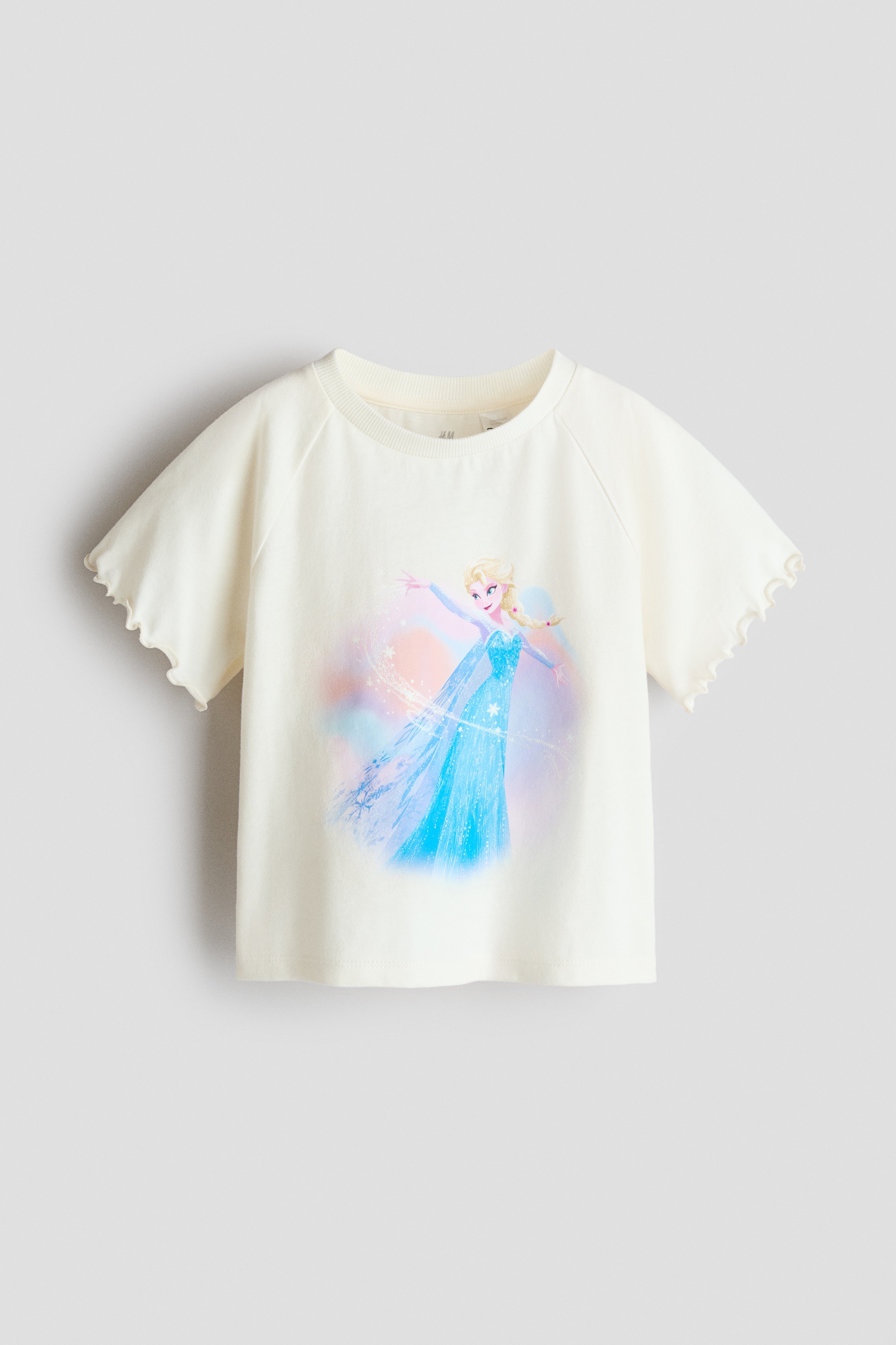 A cream-coloured cotton jersey T-shirt featuring a printed graphic of Elsa from Frozen is being worn by the model, tucked into a pastel tulle skirt. The tee features a round, rib-trimmed neckline and short raglan sleeves with wavy overlocked edges.