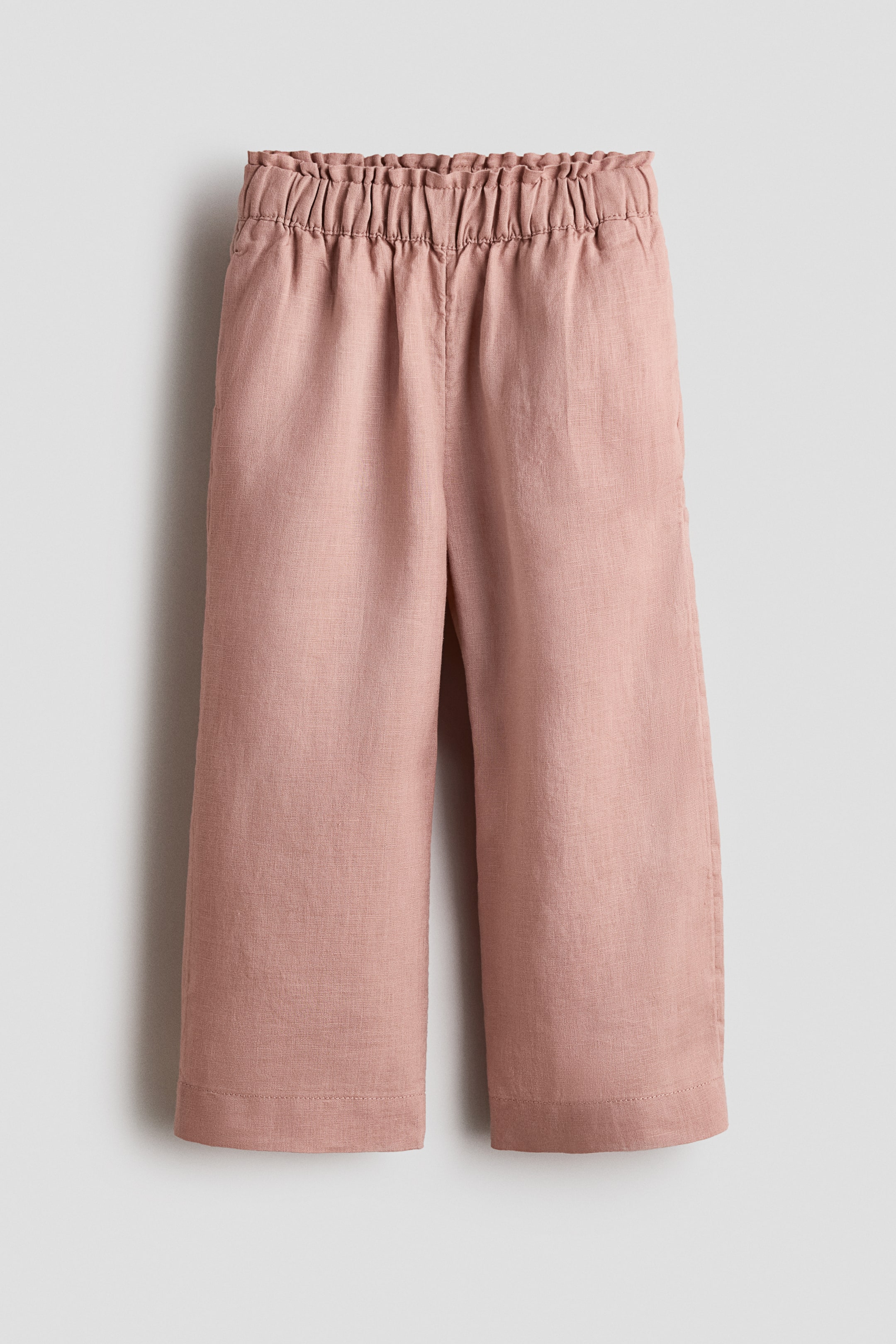 Agrandir l'image: Un pantalon rose pâle est montré de face, avec une taille haute élastiquée et froncée et une silhouette ample et raccourcie. Des poches latérales sont visibles aux hanches et le tissu présente un tissage texturé en lin.