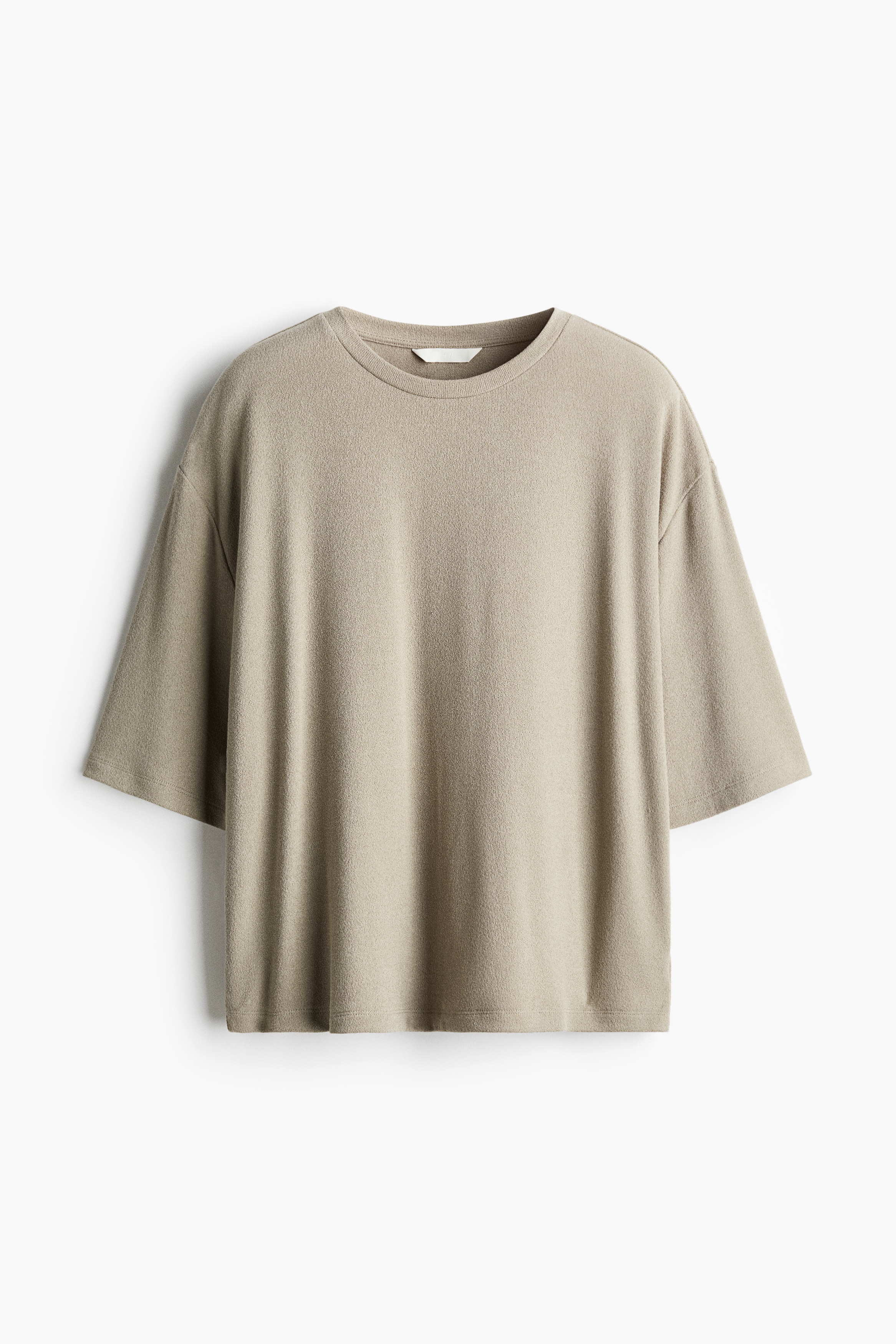 The model is wearing an oversized beige top featuring a round neckline, dropped shoulders, and 3/4-length sleeves. The top is styled with matching trousers, and the model is resting one hand in a pocket, creating a soft drape at the hem.