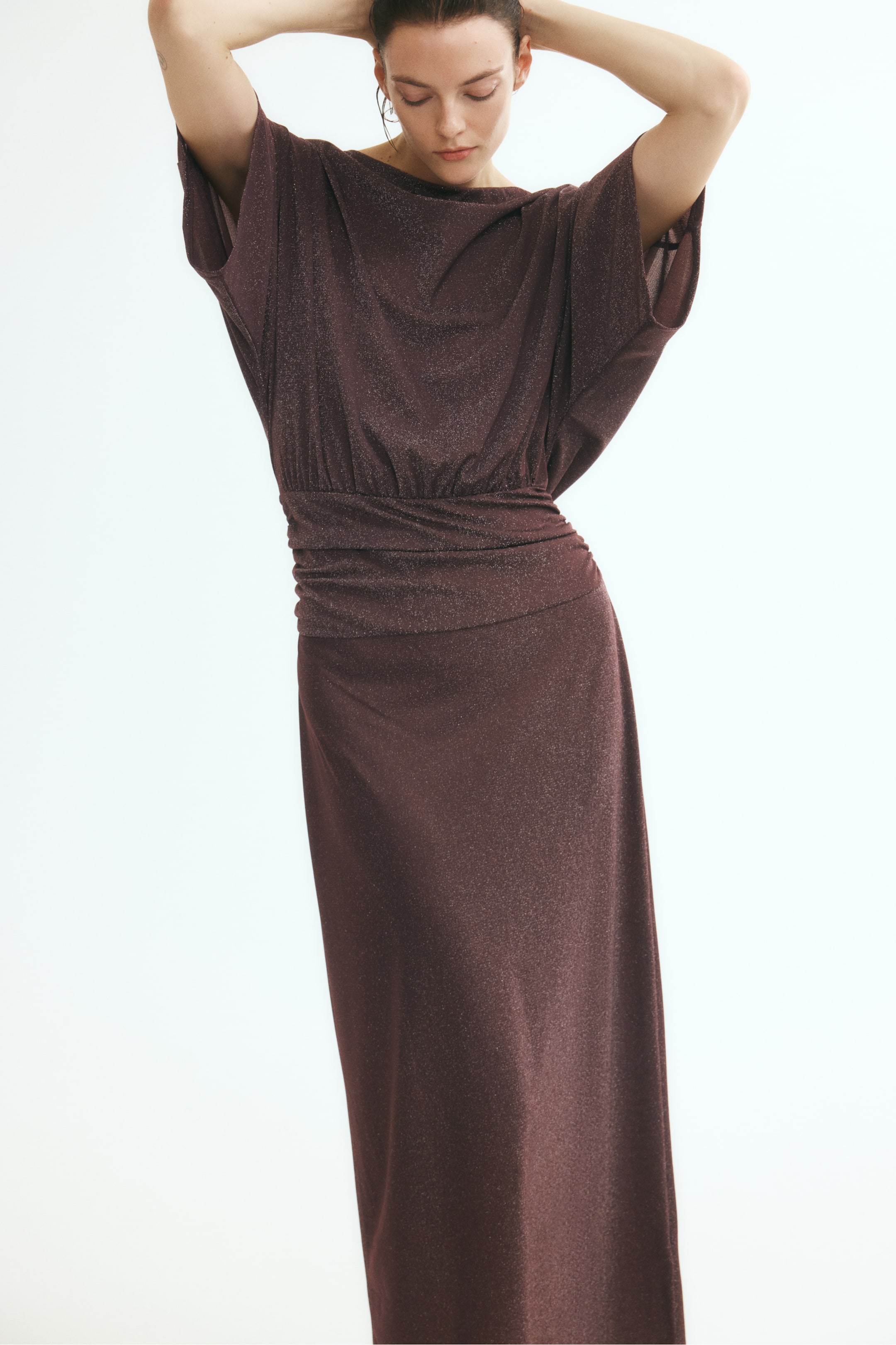 View larger image: A shimmering dark brown maxi dress is worn by the model, who is posing with her hands raised behind her head. The dress features a draped high neckline, short dolman sleeves, and a wide gathered waist panel that flows into a straight skirt.