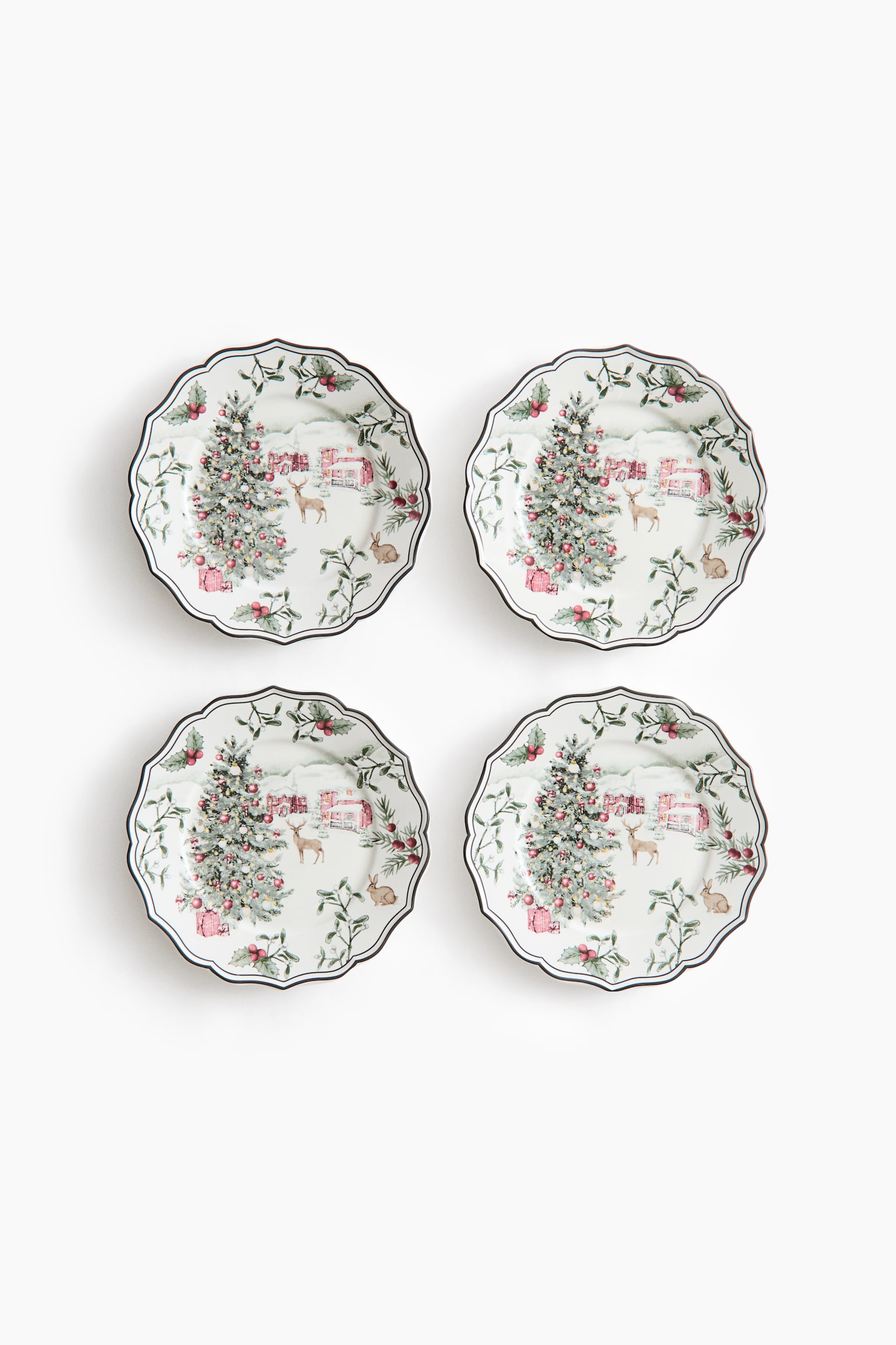 A set of four white ceramic plates is displayed from a front-facing view in a two-by-two arrangement. Each plate features a scalloped edge with a dark rim and showcases a winter scene illustration including a decorated holiday tree with gifts, a deer, and a pink cottage in a snowy landscape. - 1
