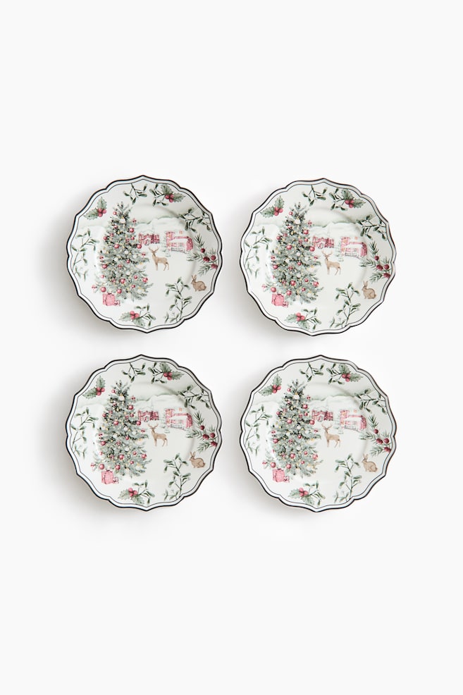 A set of four white ceramic plates is displayed from a front-facing view in a two-by-two arrangement. Each plate features a scalloped edge with a dark rim and showcases a winter scene illustration that includes a decorated holiday tree with gifts, a deer, and a pink cottage in a snowy landscape. - 1